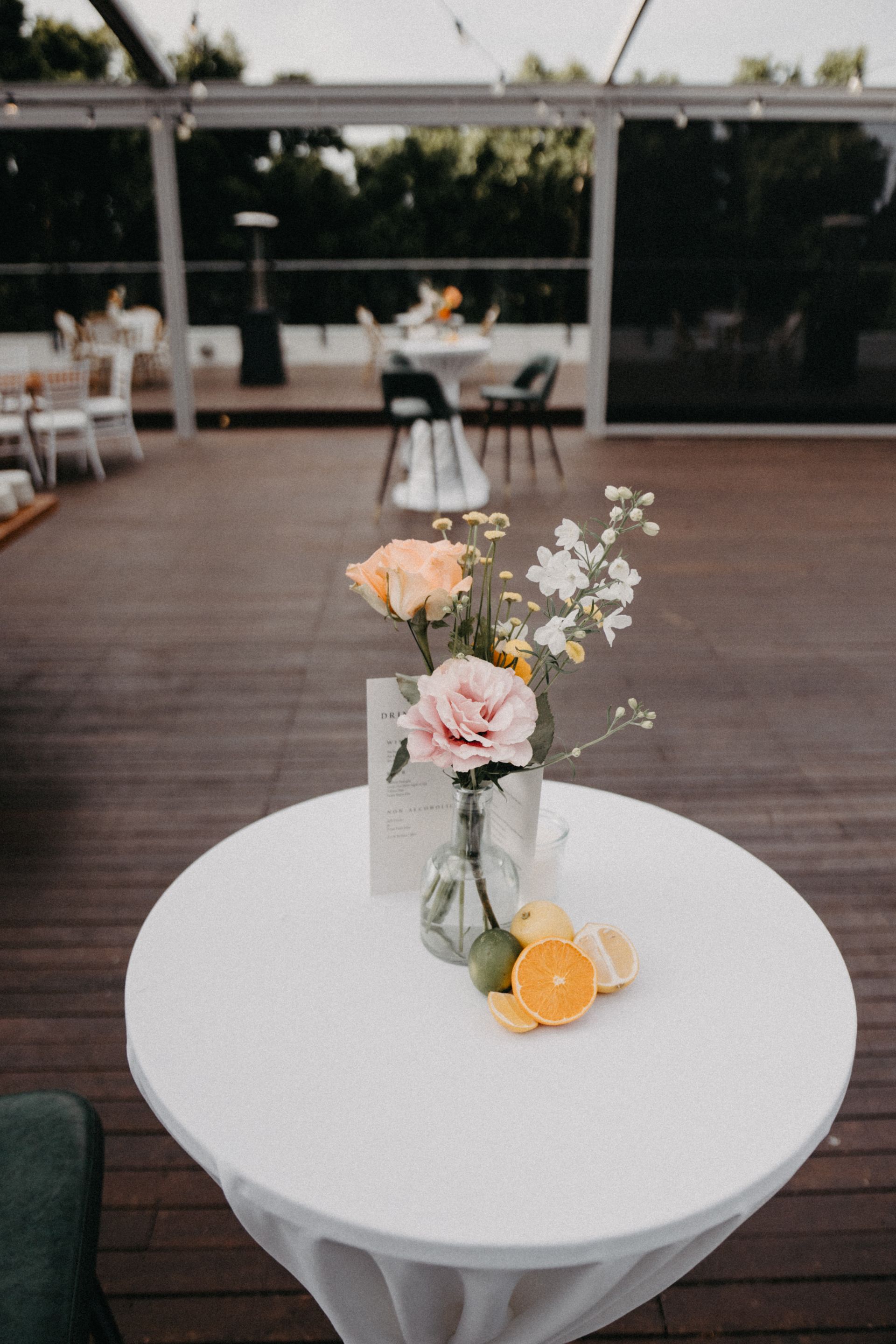 Round white table with a small vase of pink and white flowers and orange slices on a wooden patio