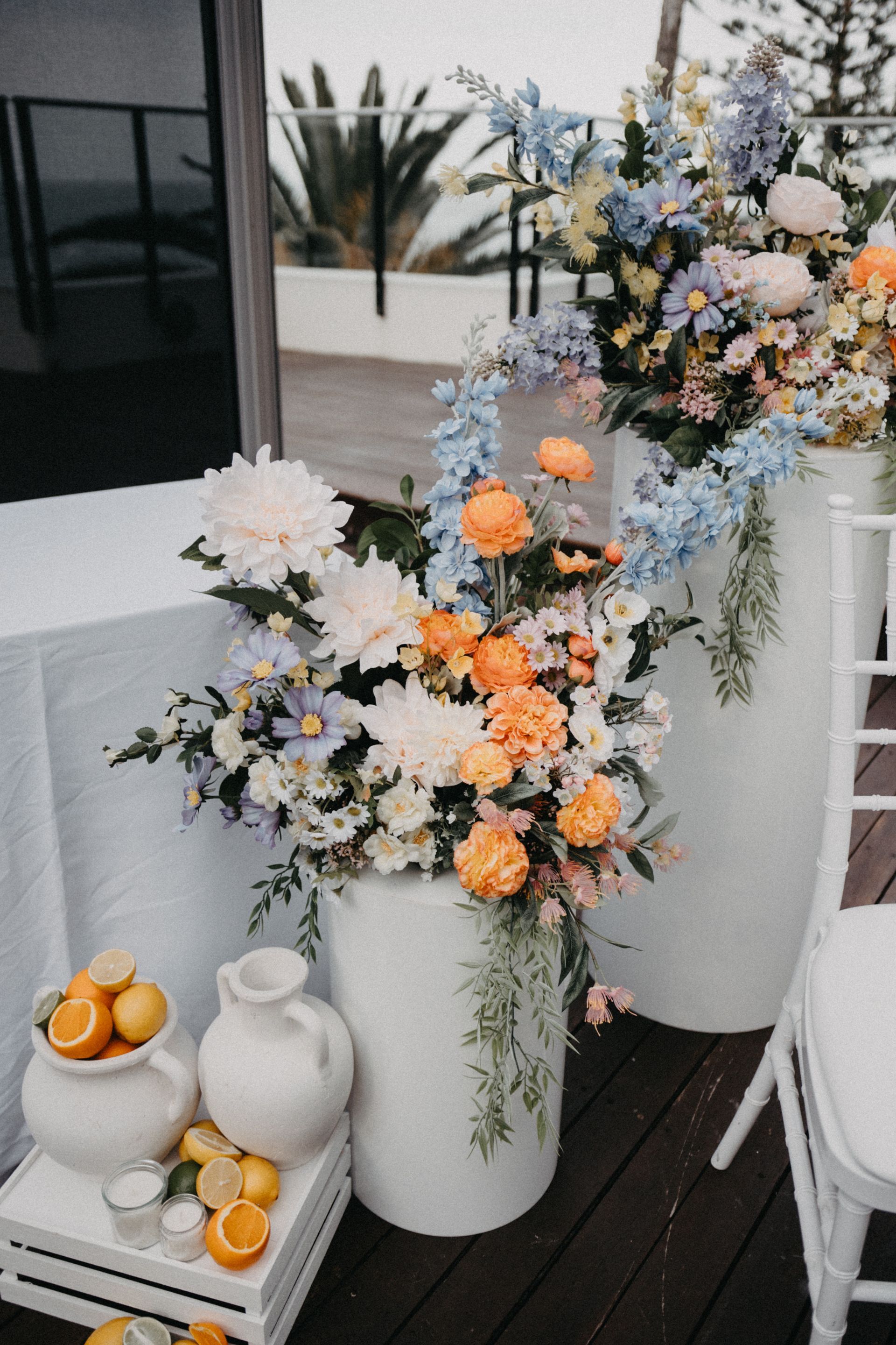 Pastel flower arrangements in white vases on a patio beside a tray of oranges and ceramic jugs