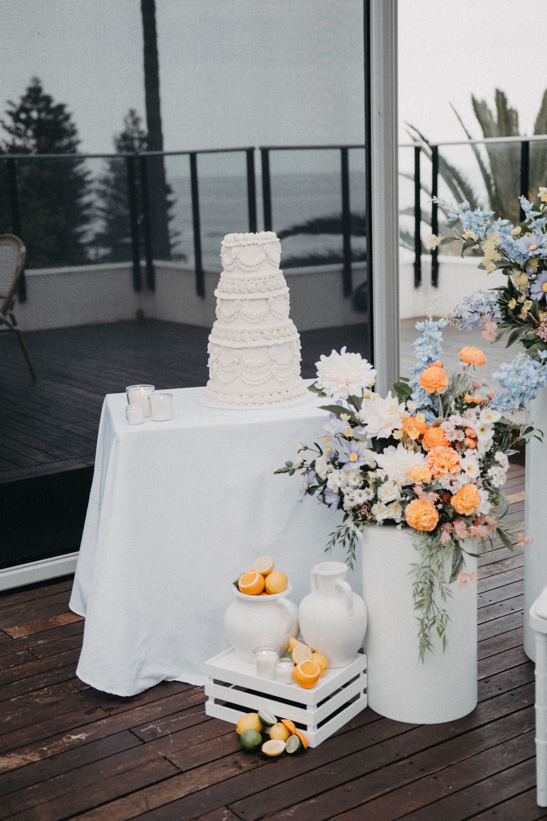 White tiered cake and floral display beside white tablecloth on an outdoor patio with lemons and candles.