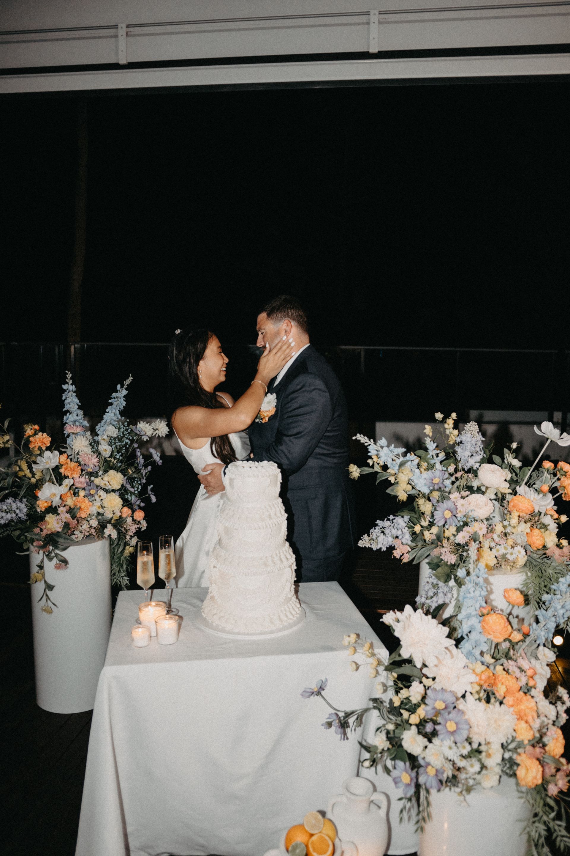 Wedding couple sharing a kiss beside a white cake and pastel floral arrangements at a reception table