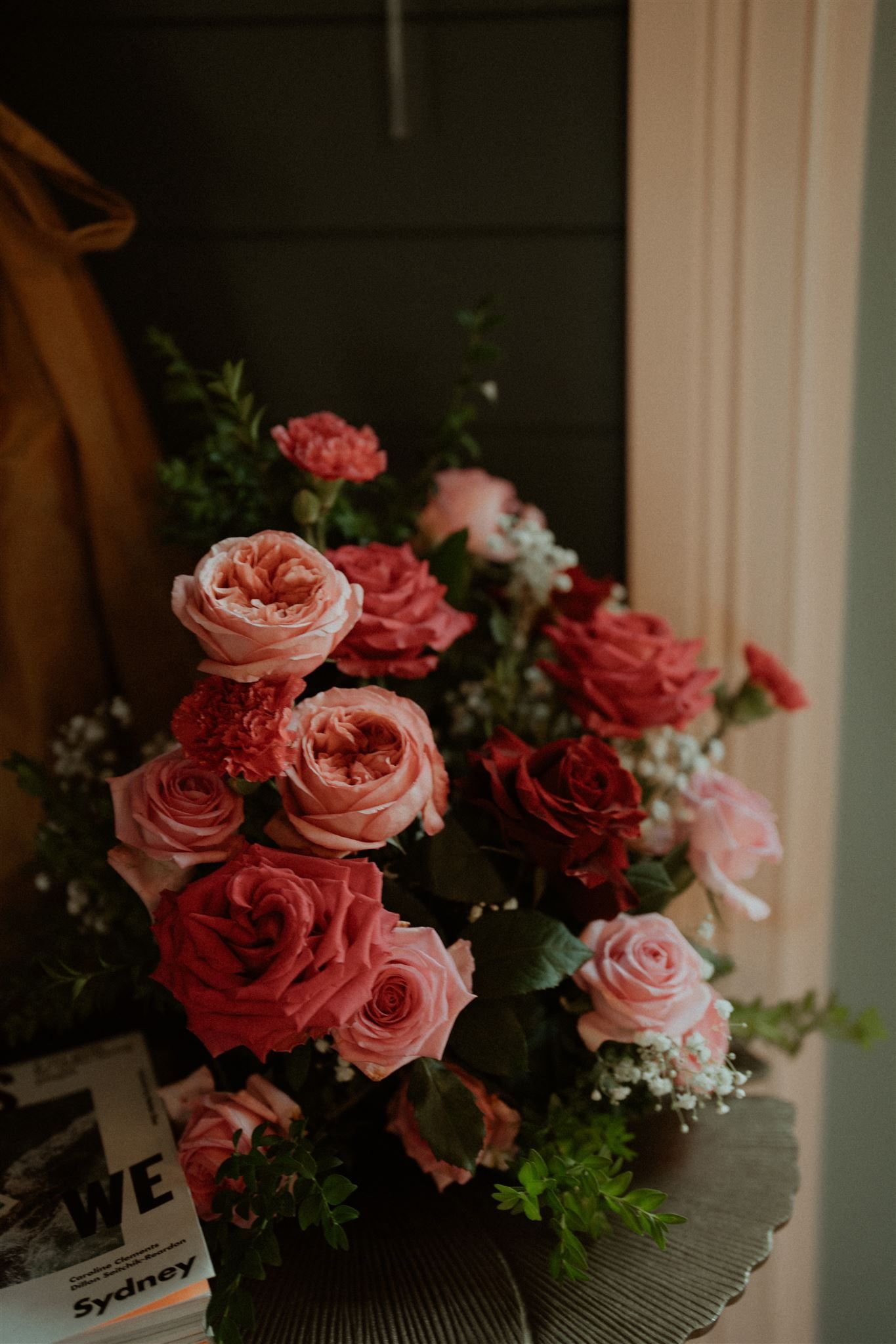Pink roses in a bouquet on a table by a window, with dark green foliage and soft indoor light