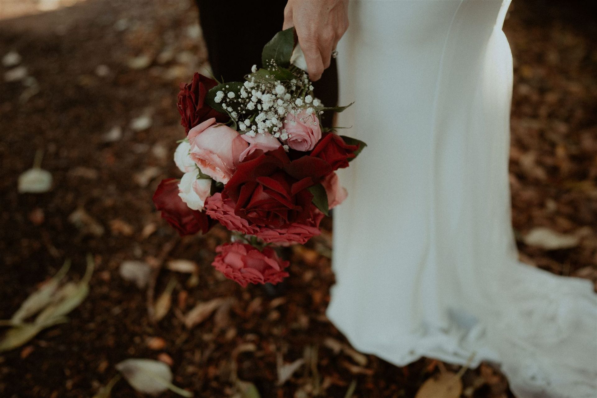 Bride holding a red and white bouquet beside a white wedding dress outdoors on fallen leaves