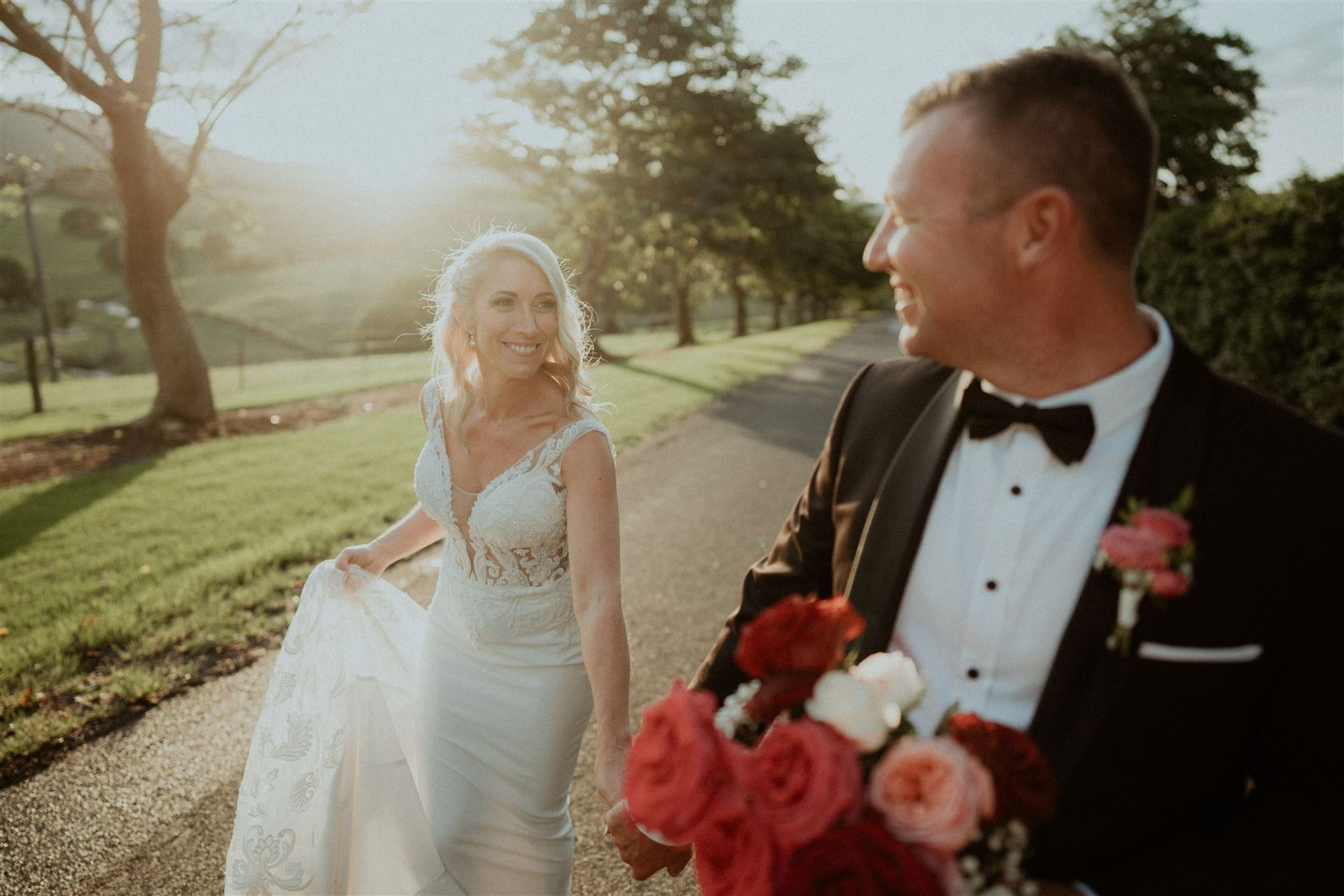 Bride and groom walking outdoors at sunset, smiling and holding red and white flowers
