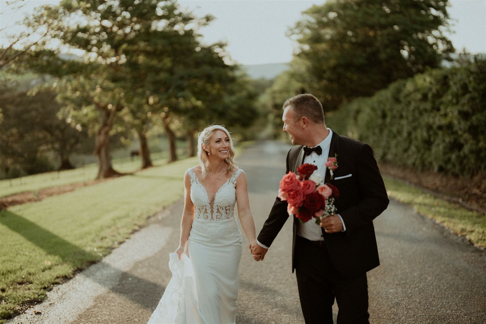 Bride and groom walking hand in hand on a tree-lined road, smiling and holding a bouquet.