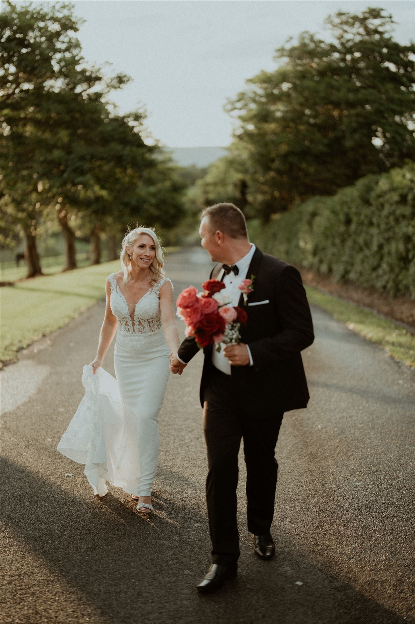 Bride and groom walking down a country road, smiling and holding red flowers.