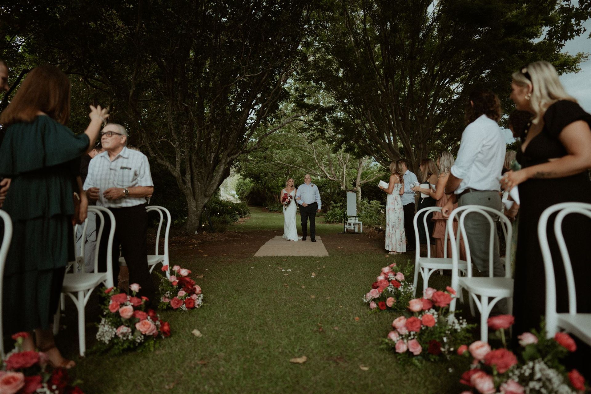 Outdoor wedding aisle with white chairs, floral arrangements, and guests under trees