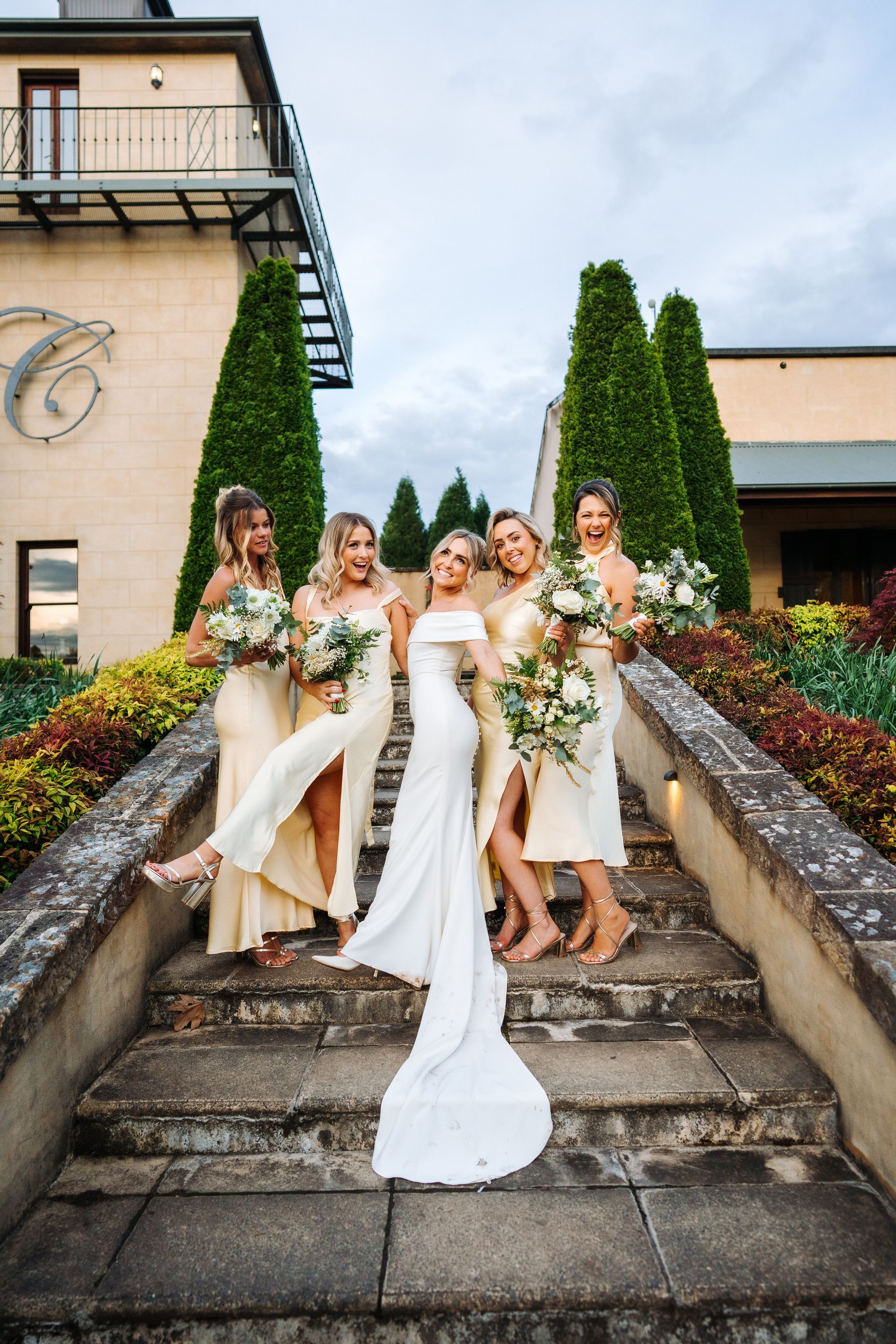 Bride and bridesmaids on steps, posing for photo. Bride in white gown, bridesmaids in yellow dresses, outside.