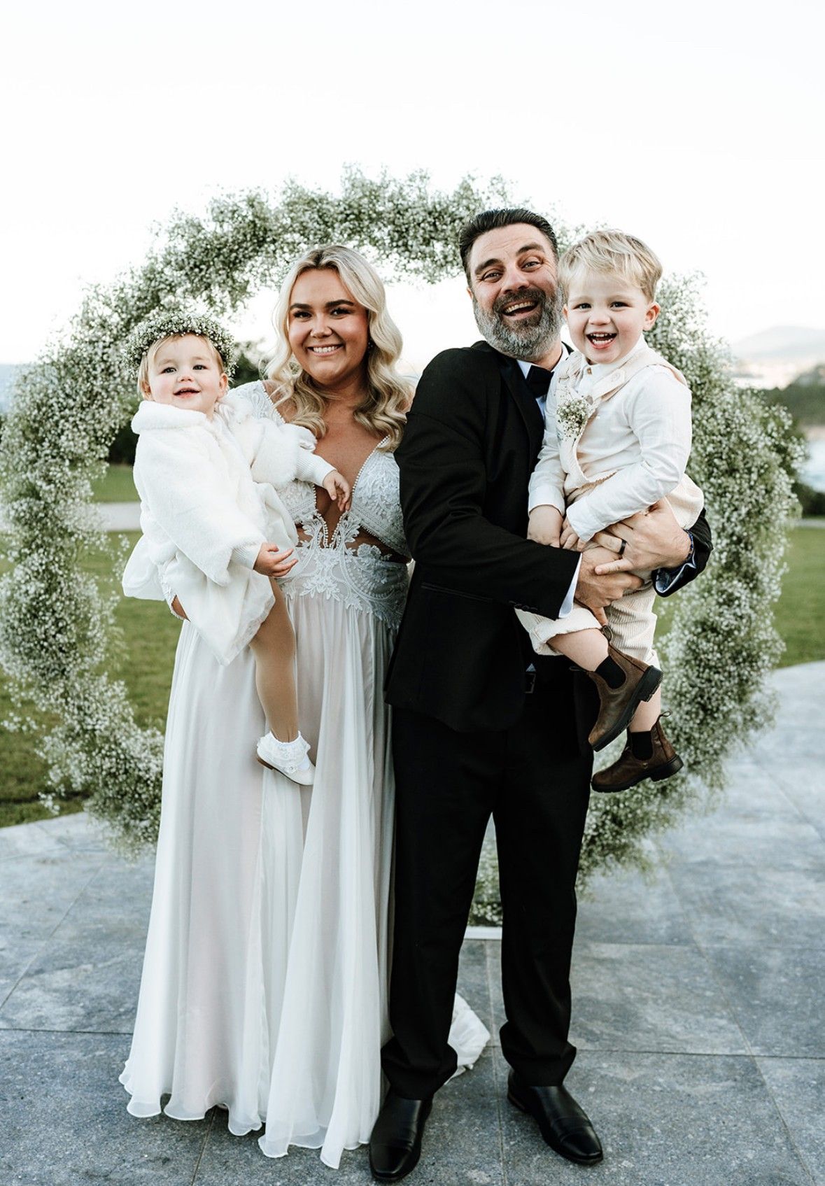 Family of four posing outdoors in formal white and black outfits by a floral arch