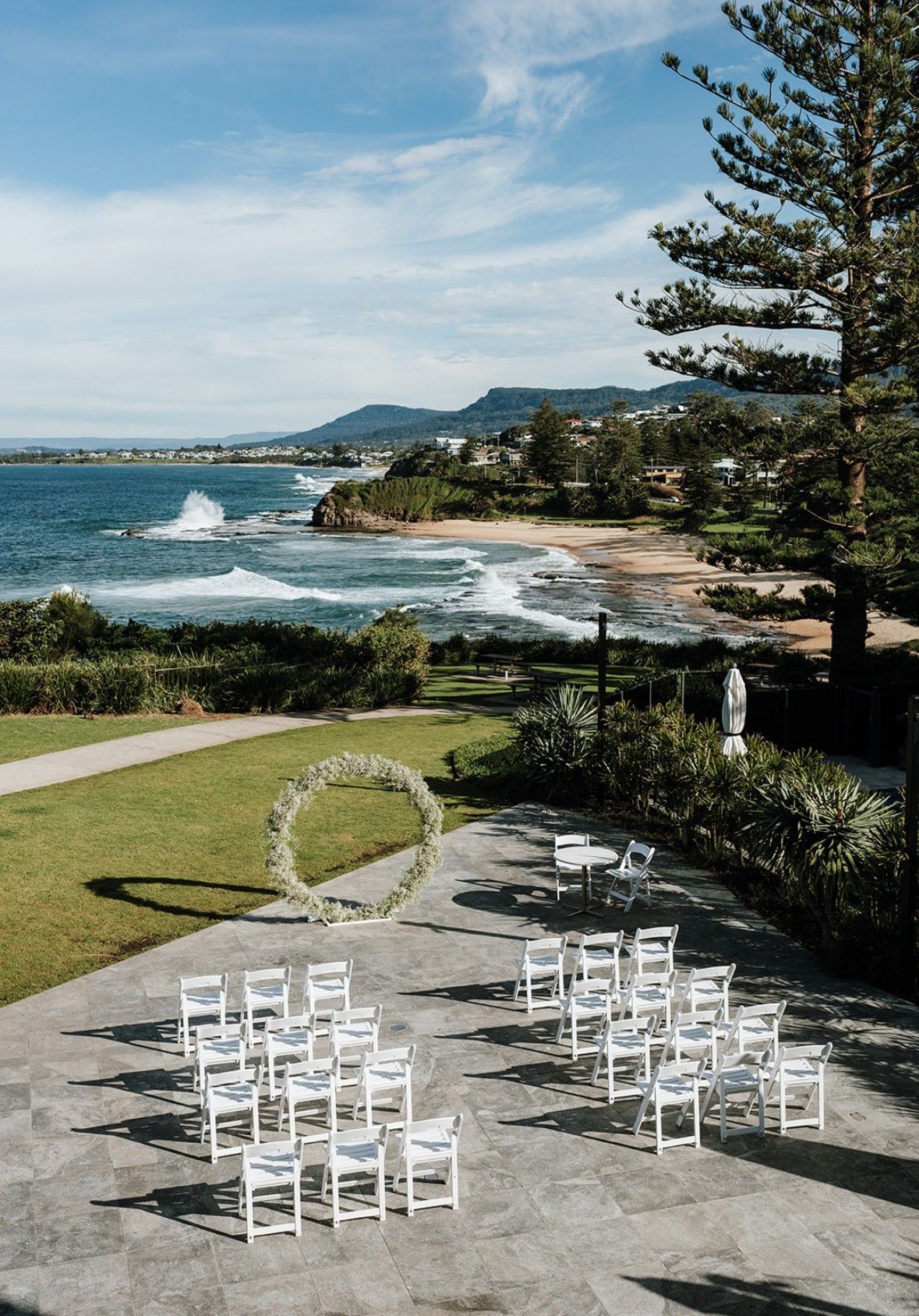 Outdoor wedding setup with white chairs on a seaside lawn and a floral arch overlooking the ocean