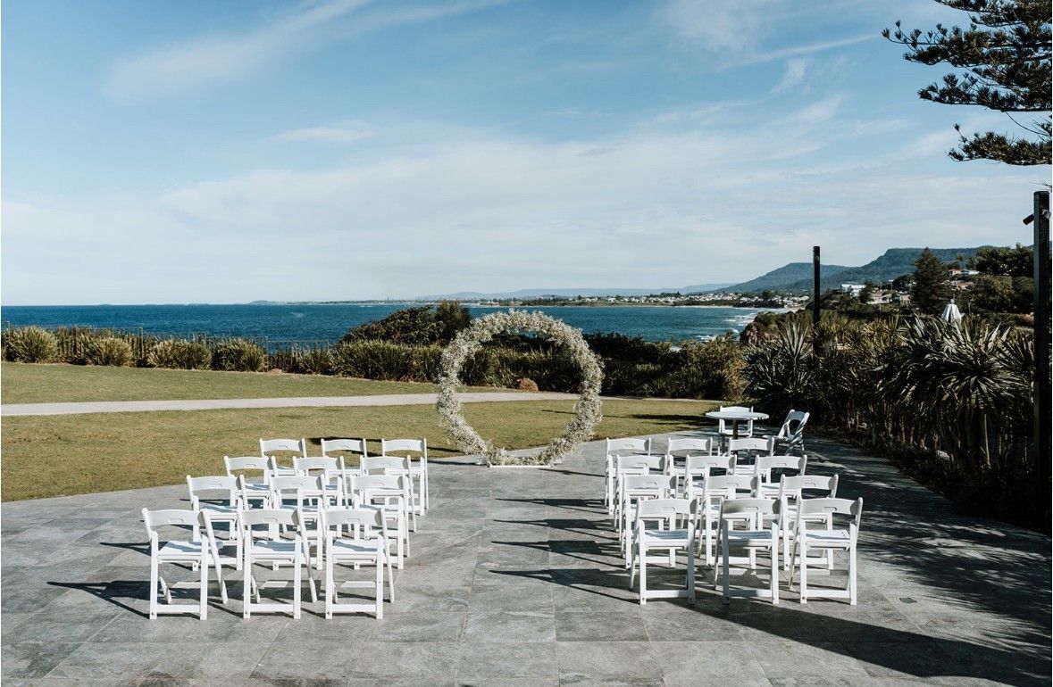 White chairs arranged for an outdoor wedding ceremony facing a floral arch by the sea