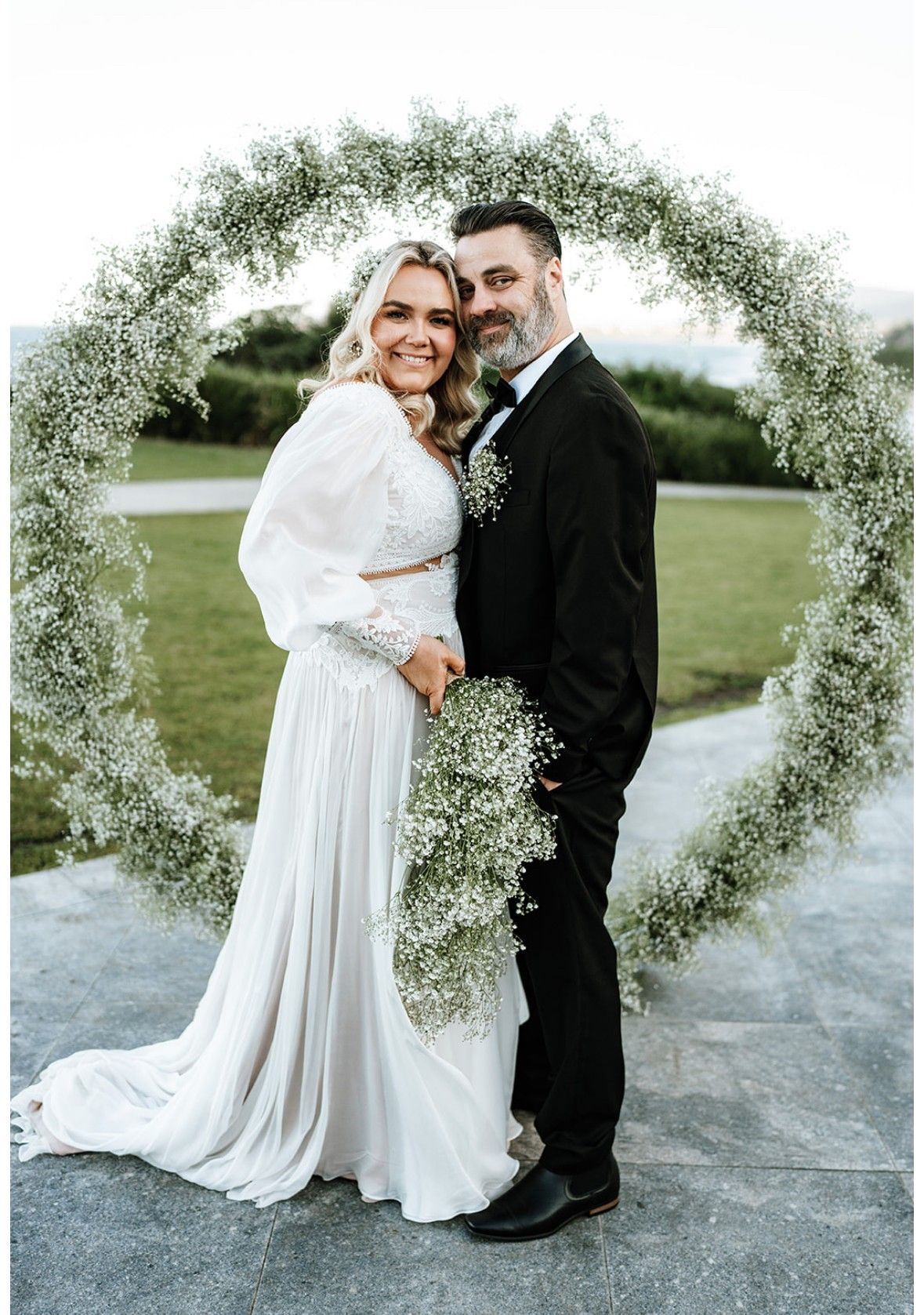 Wedding couple posing under a white floral arch outdoors on a lawn