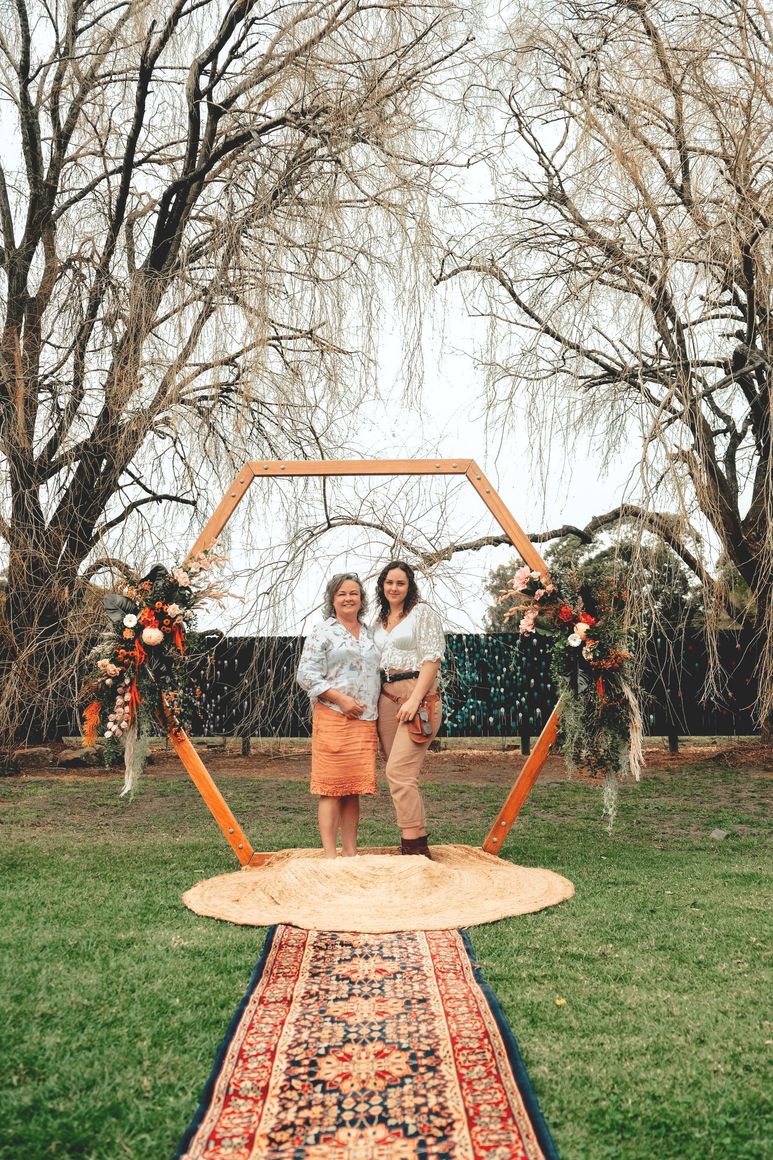 Two people standing on a rug under an orange hexagon arch in a grassy outdoor setting with trees and flowers.