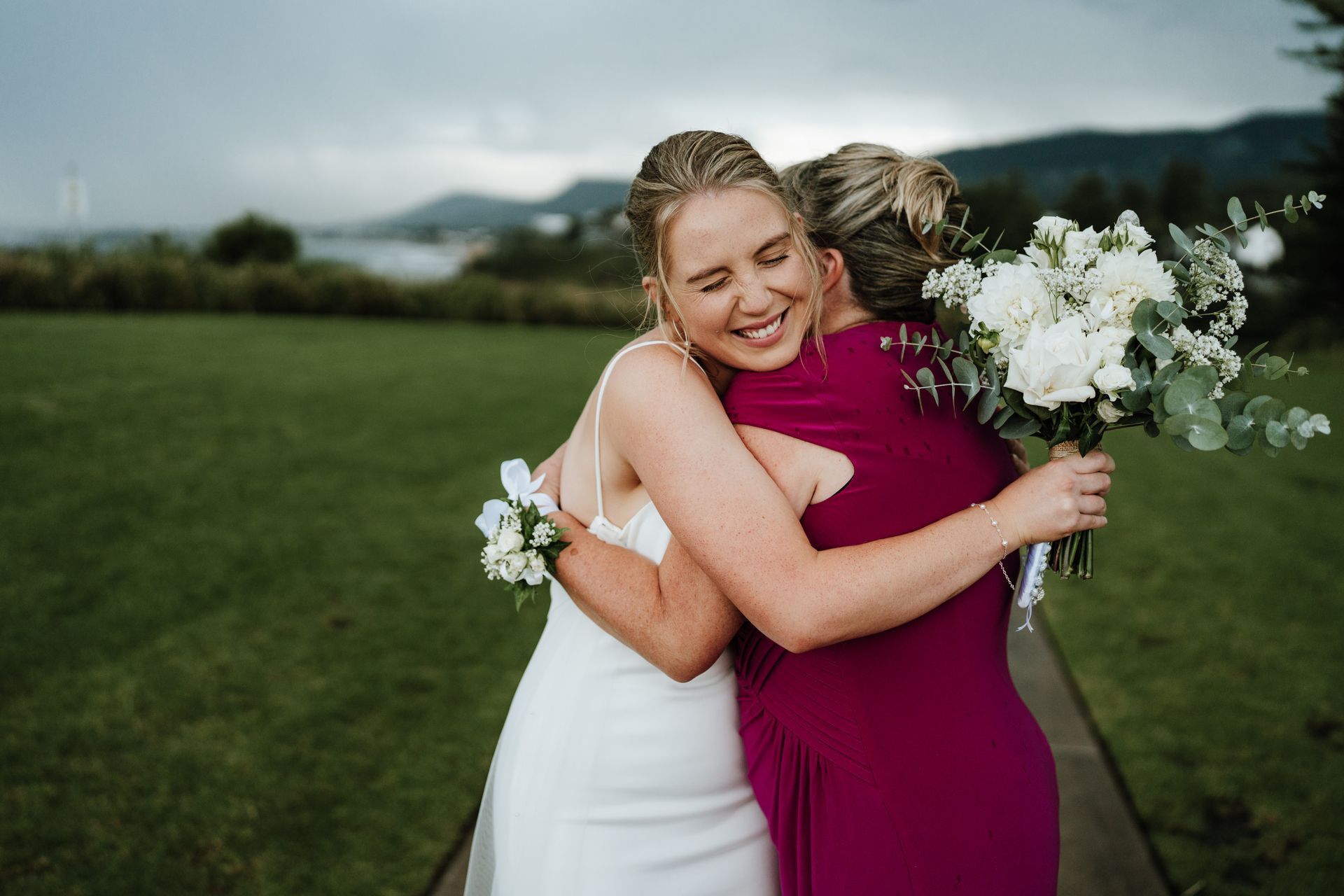 Two people in white and magenta dresses hug outdoors on a grassy field, one holding a bouquet