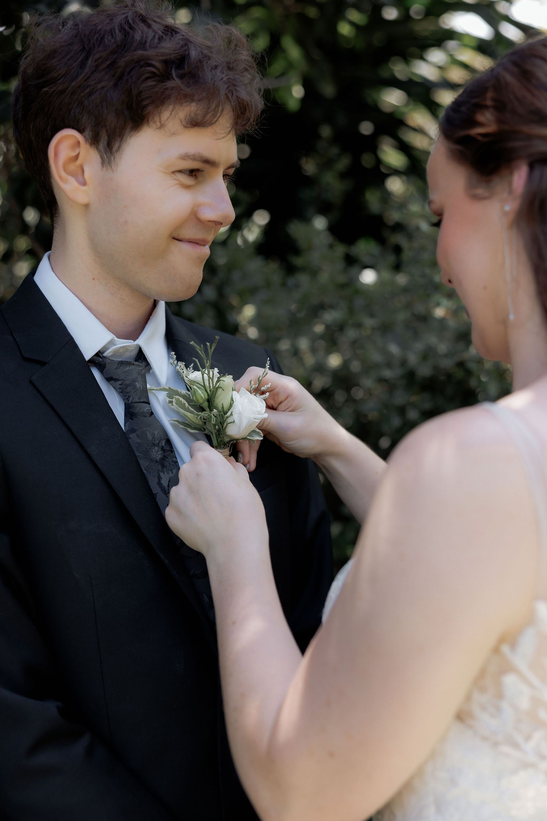 Wedding couple adjusting a boutonniere outdoors, with a smiling groom in a dark suit.