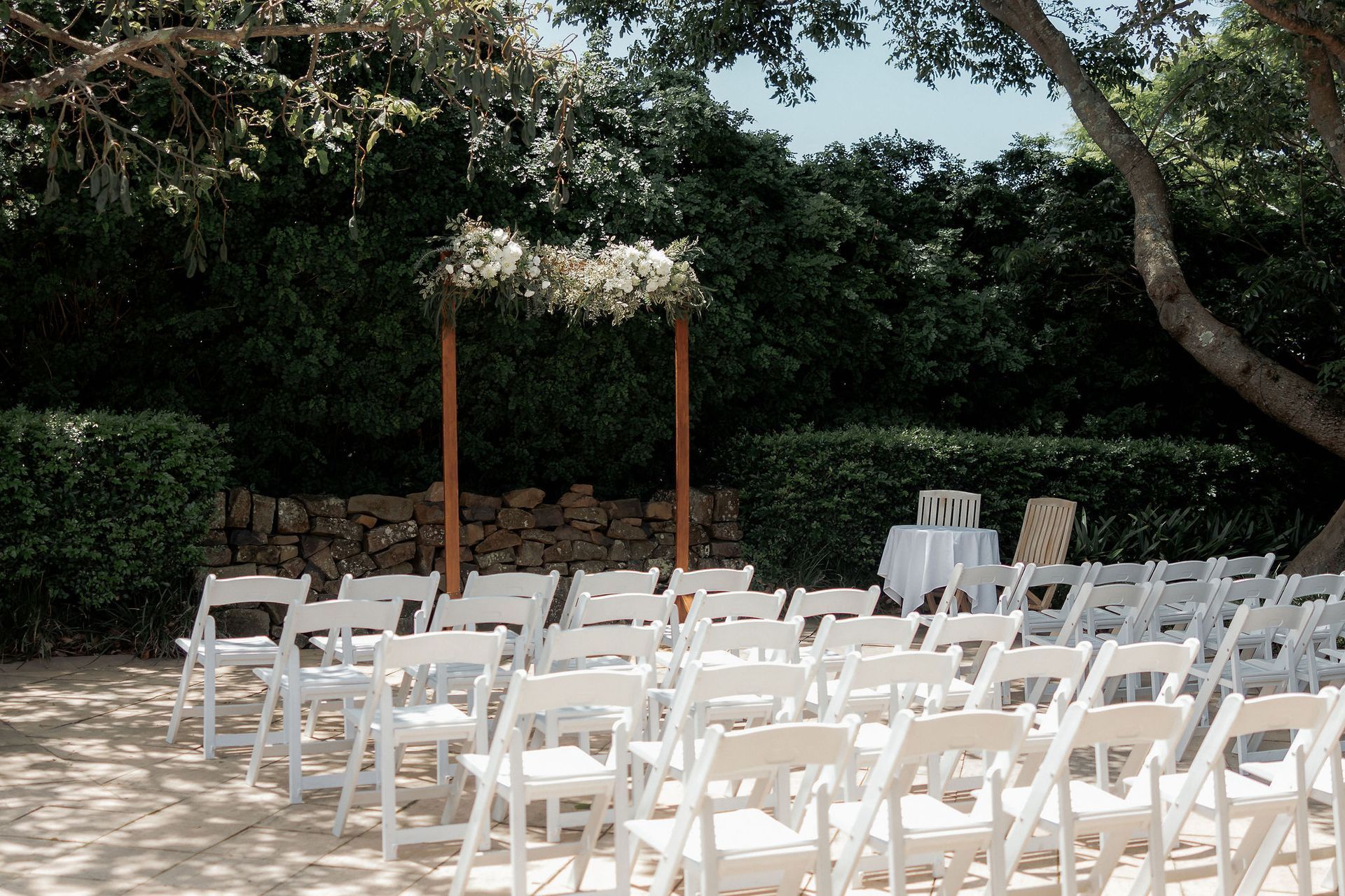 Outdoor event setup with rows of white chairs under trees and a wooden arch on a sandy lawn