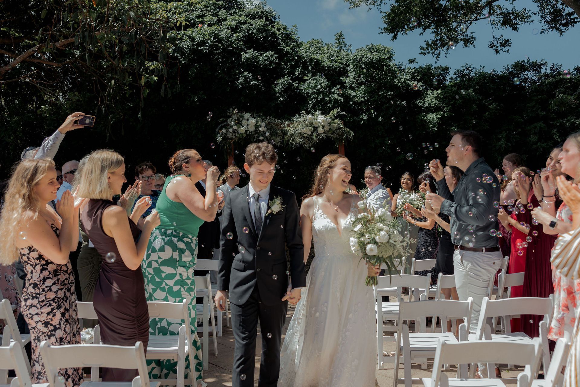 Outdoor wedding aisle with guests applauding as the bride and groom walk between white chairs
