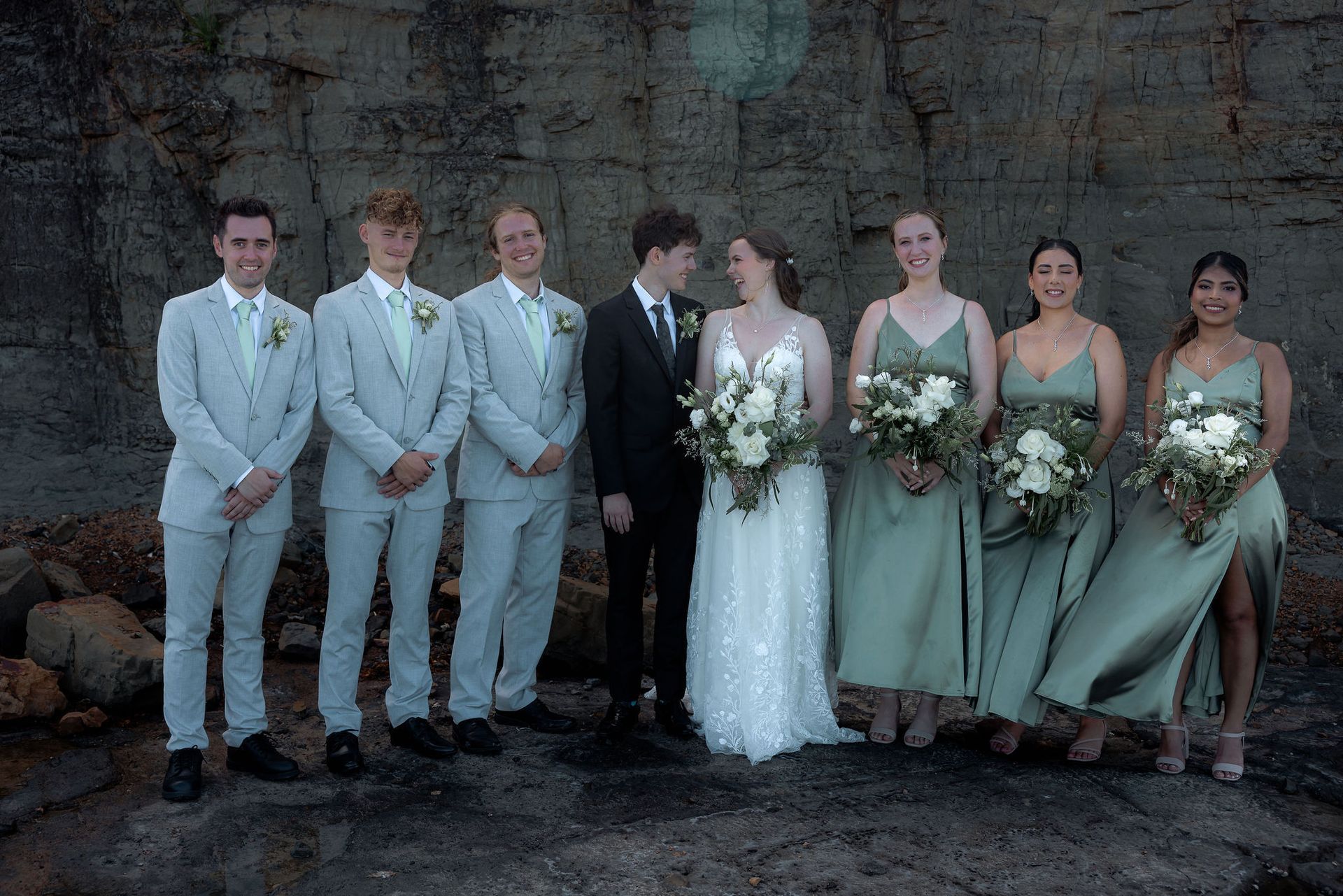 Wedding party posing outdoors, with bridesmaids in sage dresses and groomsmen in light gray suits.