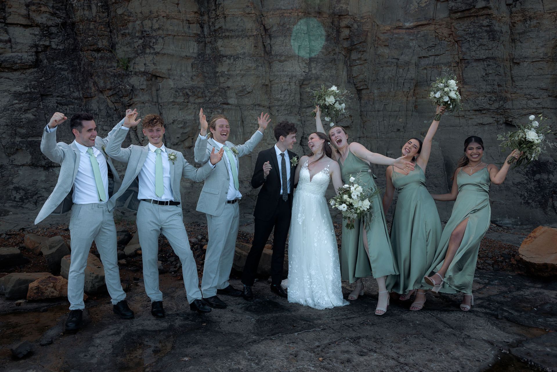 Wedding party posing on rocks by a cliff, holding bouquets and raising arms in celebration.