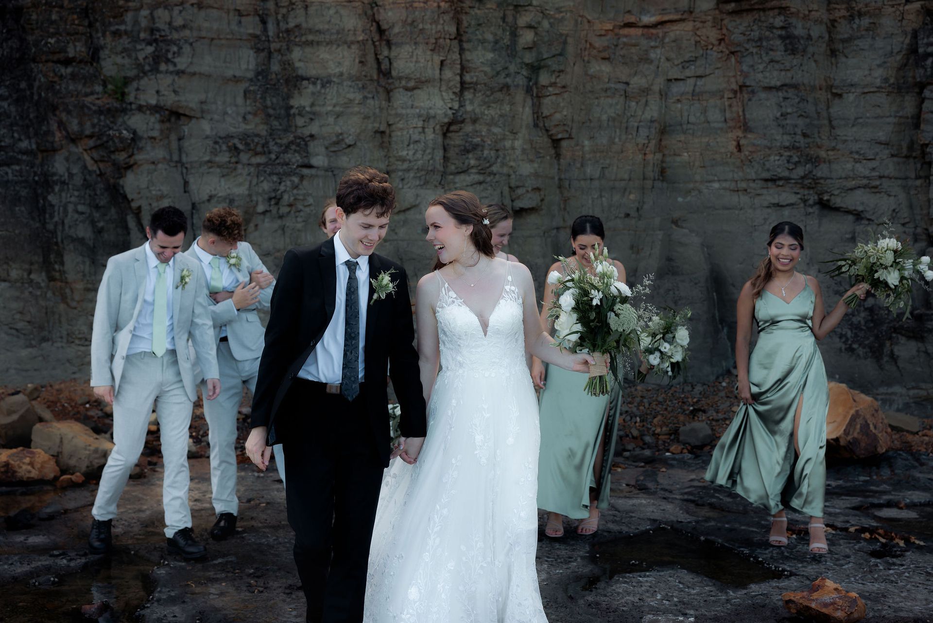 Wedding party posing outdoors against a rocky wall; bride in white gown and groom in black suit at center.