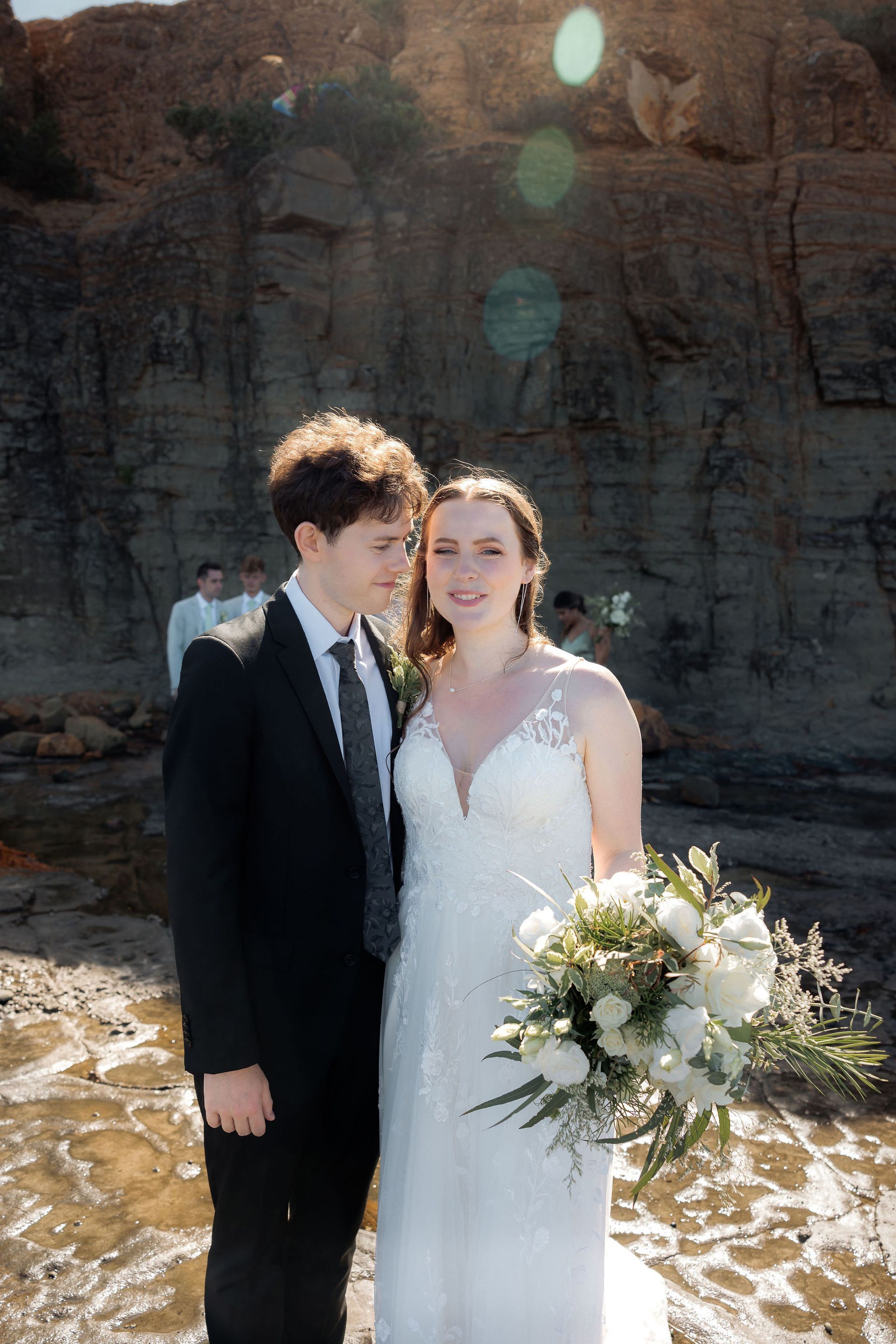 Bride and groom in black and white wedding attire standing by rocky water with sun flare