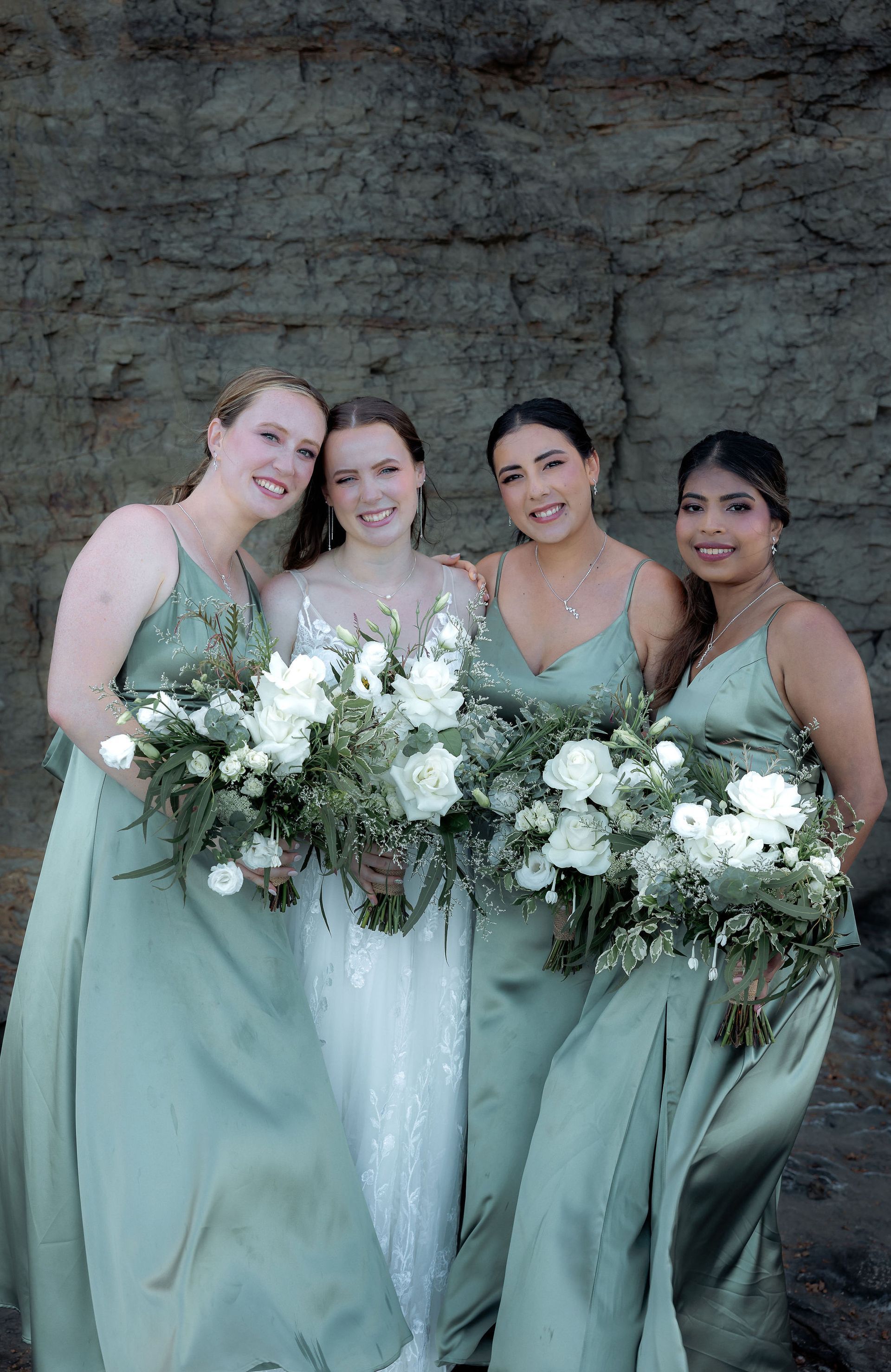 Four women in sage dresses holding white flower bouquets against a stone wall