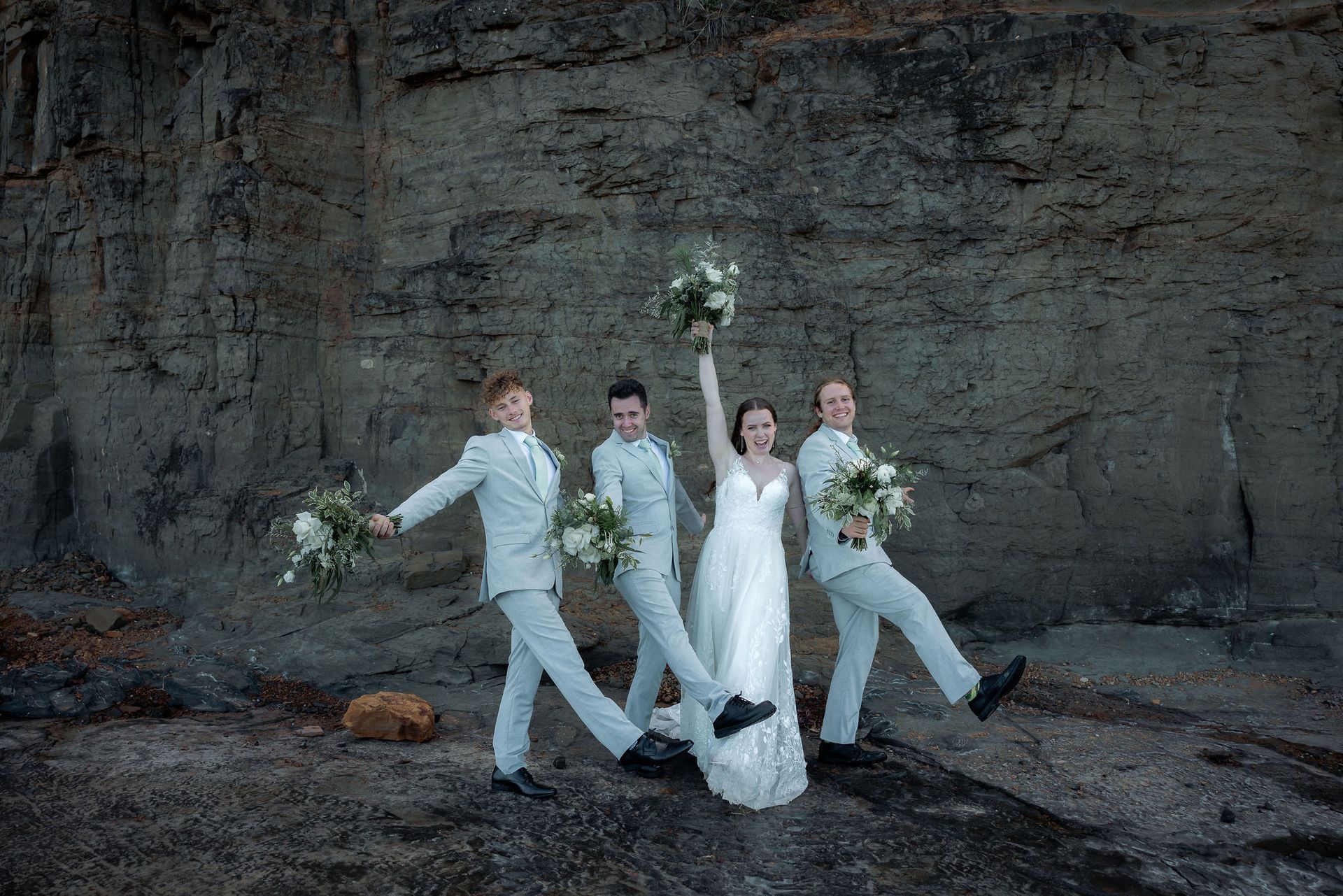 Four wedding party members pose on a rocky beach cave, holding bouquets and raising one arm.
