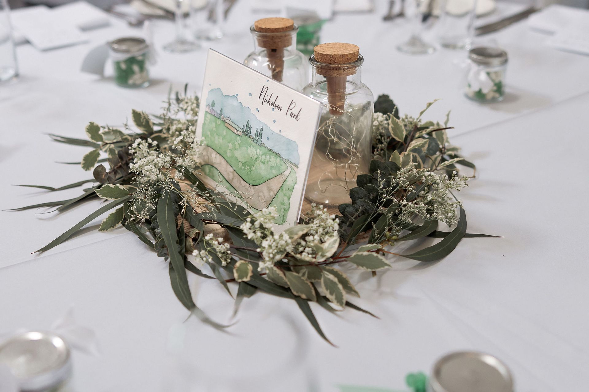 Floral table centerpiece with greenery, dried flowers, and glass bottles around a small printed card on a white tablecloth