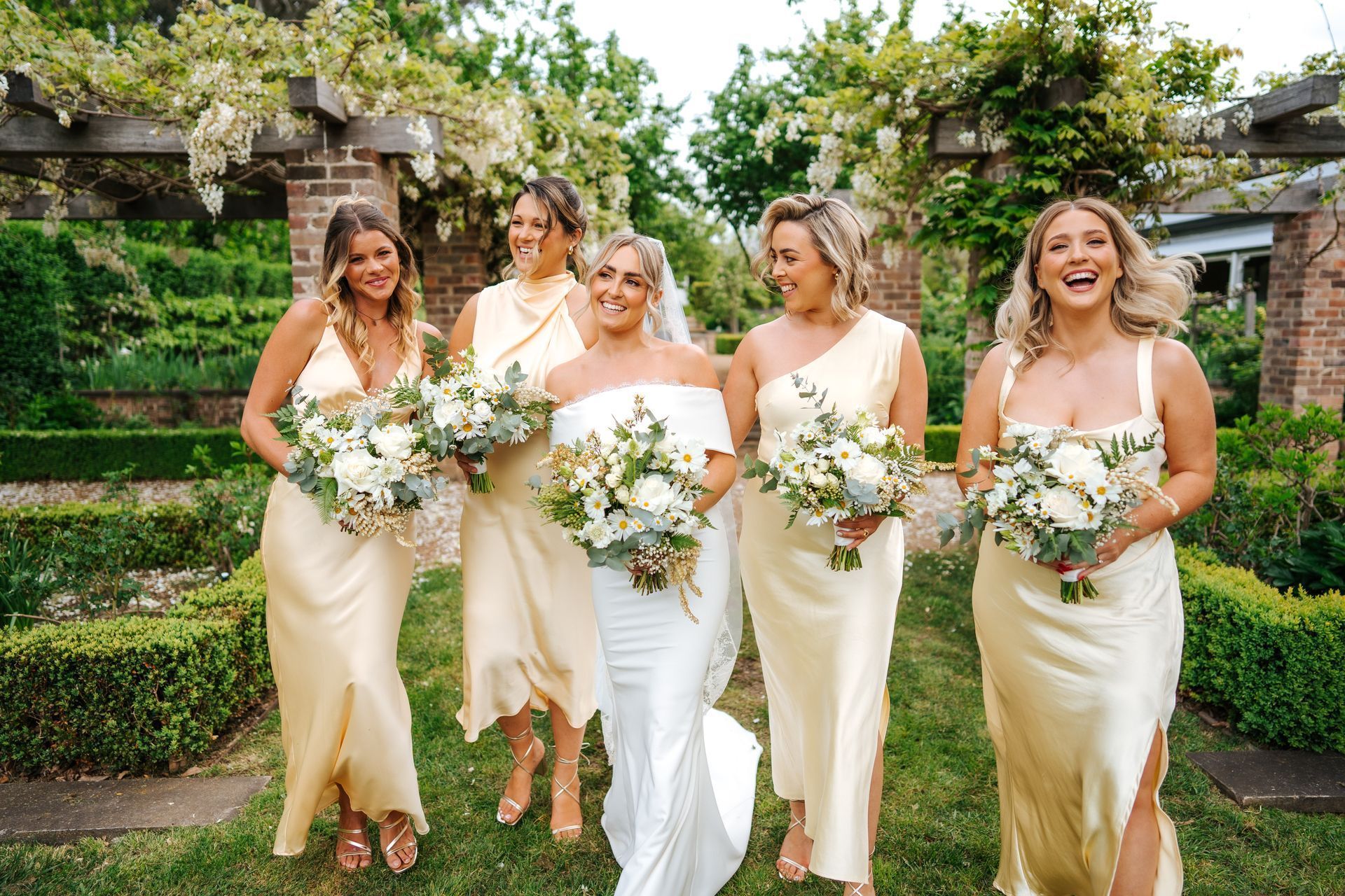 Bride and bridesmaids in yellow dresses, laughing in a garden, holding bouquets.