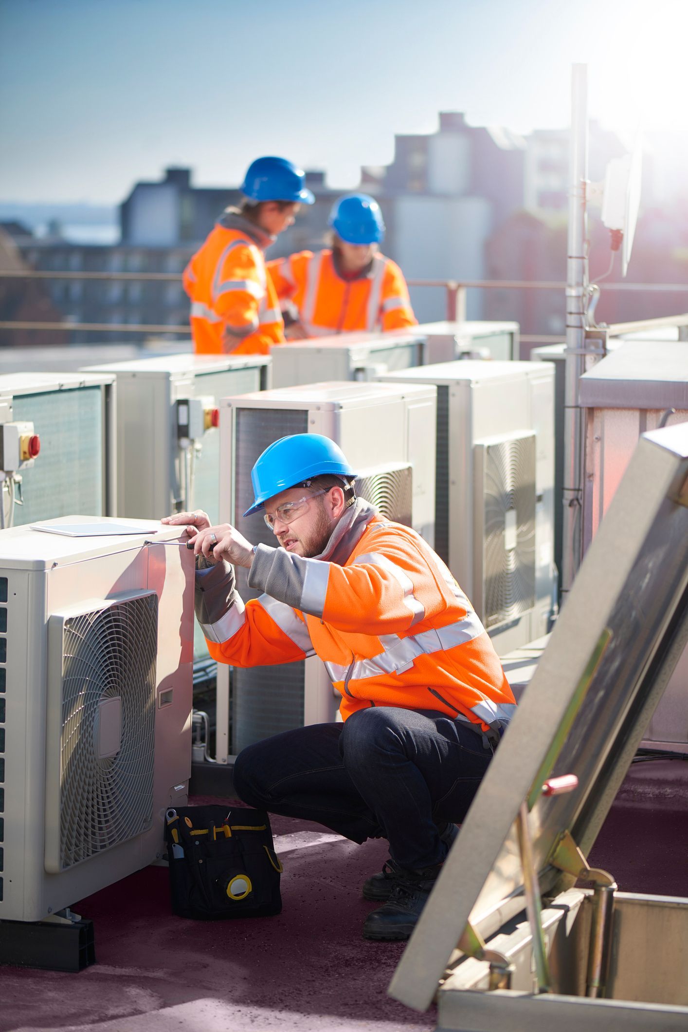 HVAC technician in orange vest and blue hardhat repairs unit on rooftop. Two colleagues in background.