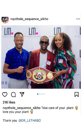 Three people posing with a boxing championship belt. One man is in a suit, holding the belt, and two people are beside him.