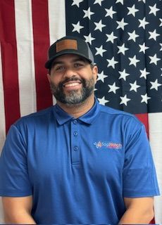 Smiling man in blue polo and cap standing before an American flag backdrop