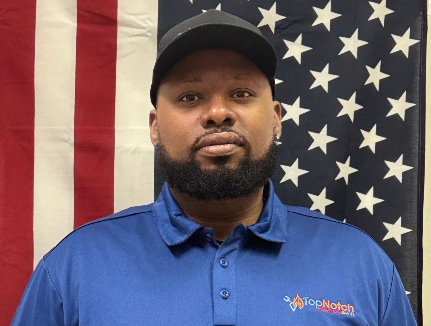 Man in a blue polo and black cap standing before an American flag backdrop
