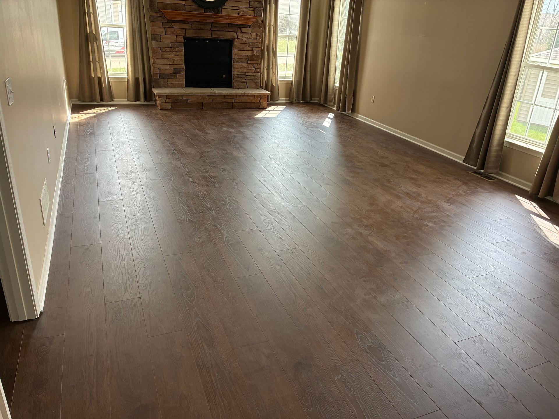 Empty living room with dark wood floors, beige walls, and a stone fireplace at the far end.