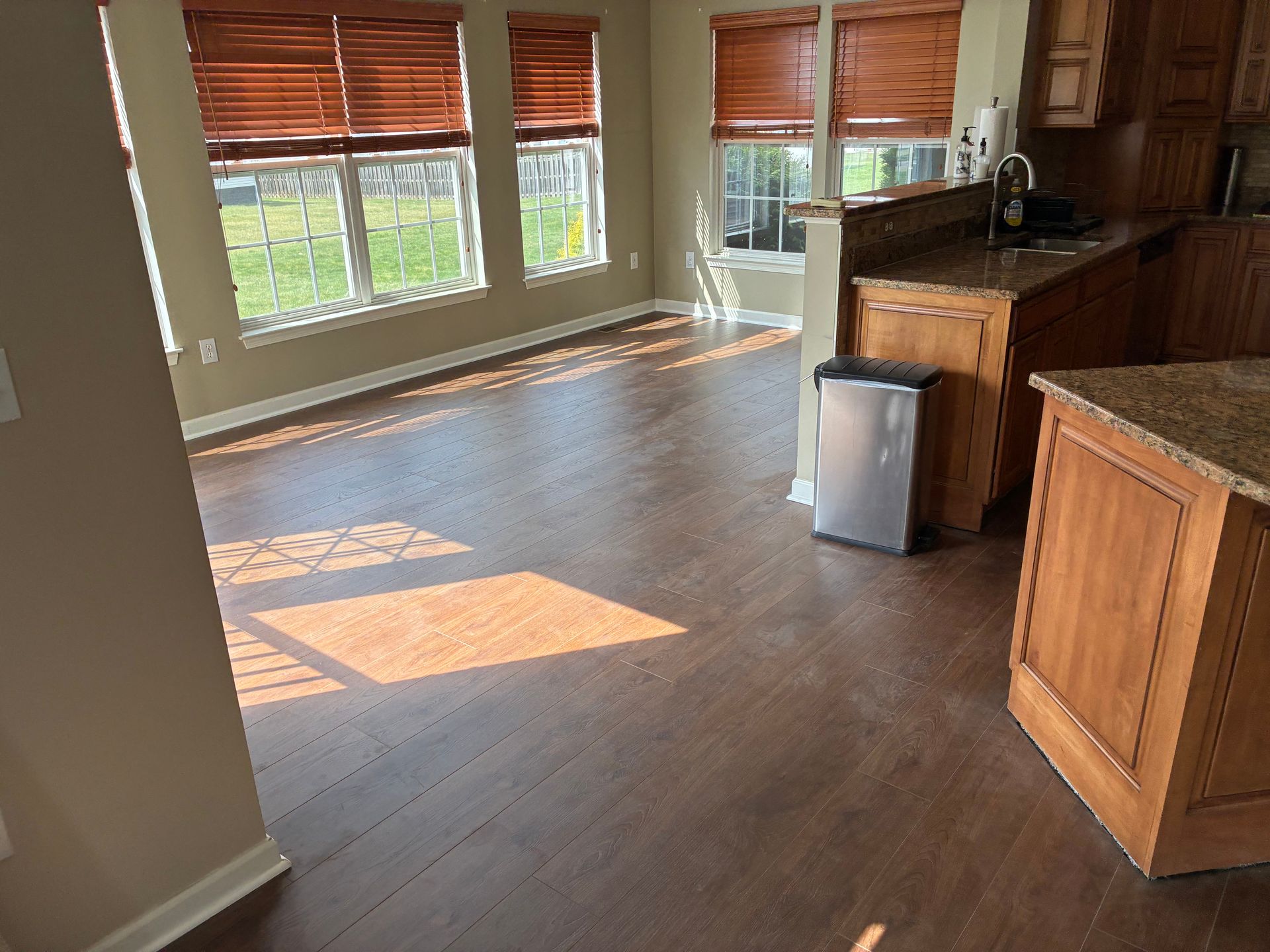 Empty kitchen with hardwood floors, wooden cabinets, and sunlight streaming through window blinds.