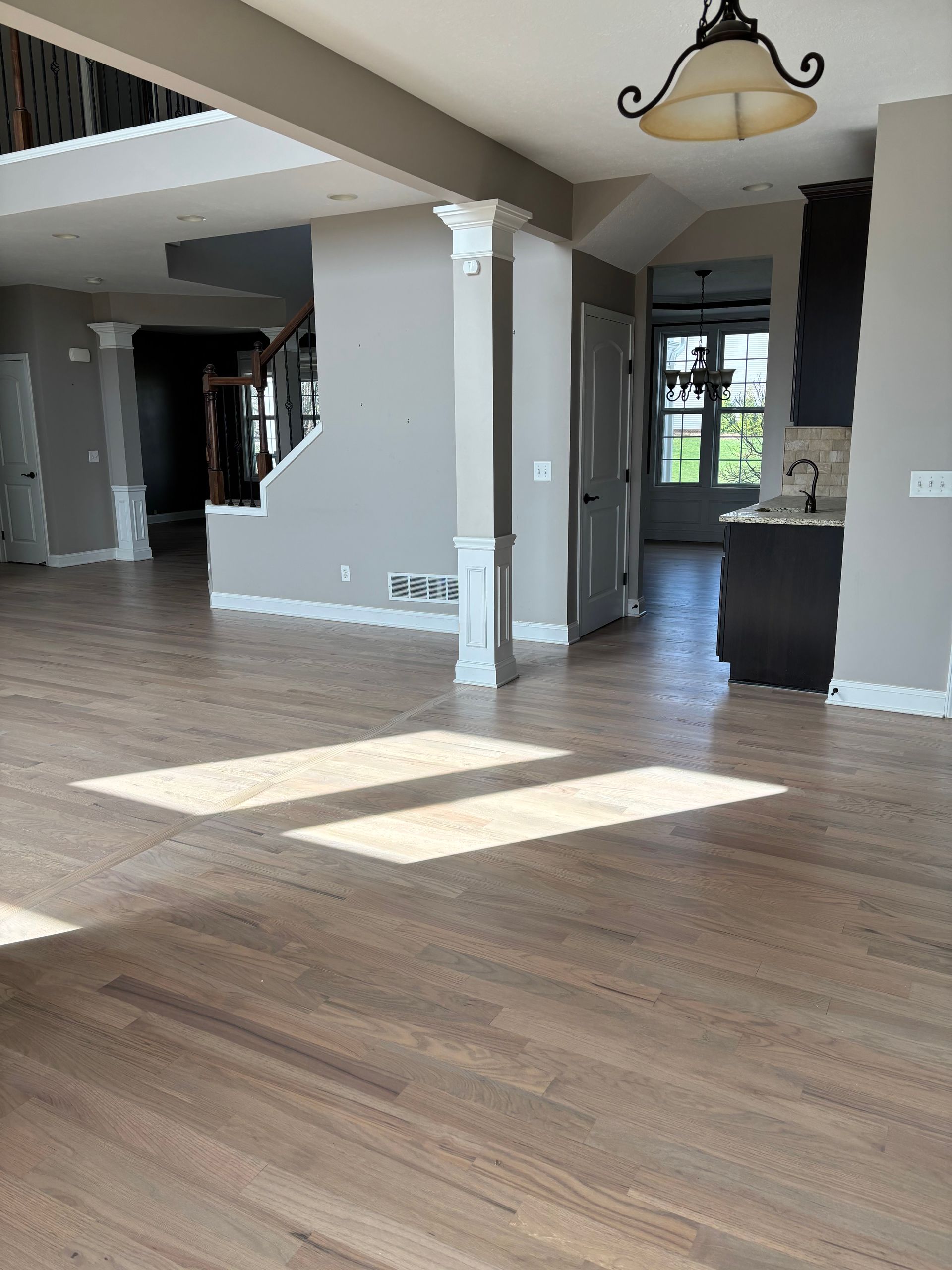 Spacious empty living room with light wood floors, gray walls, and sunlight streaming through windows.