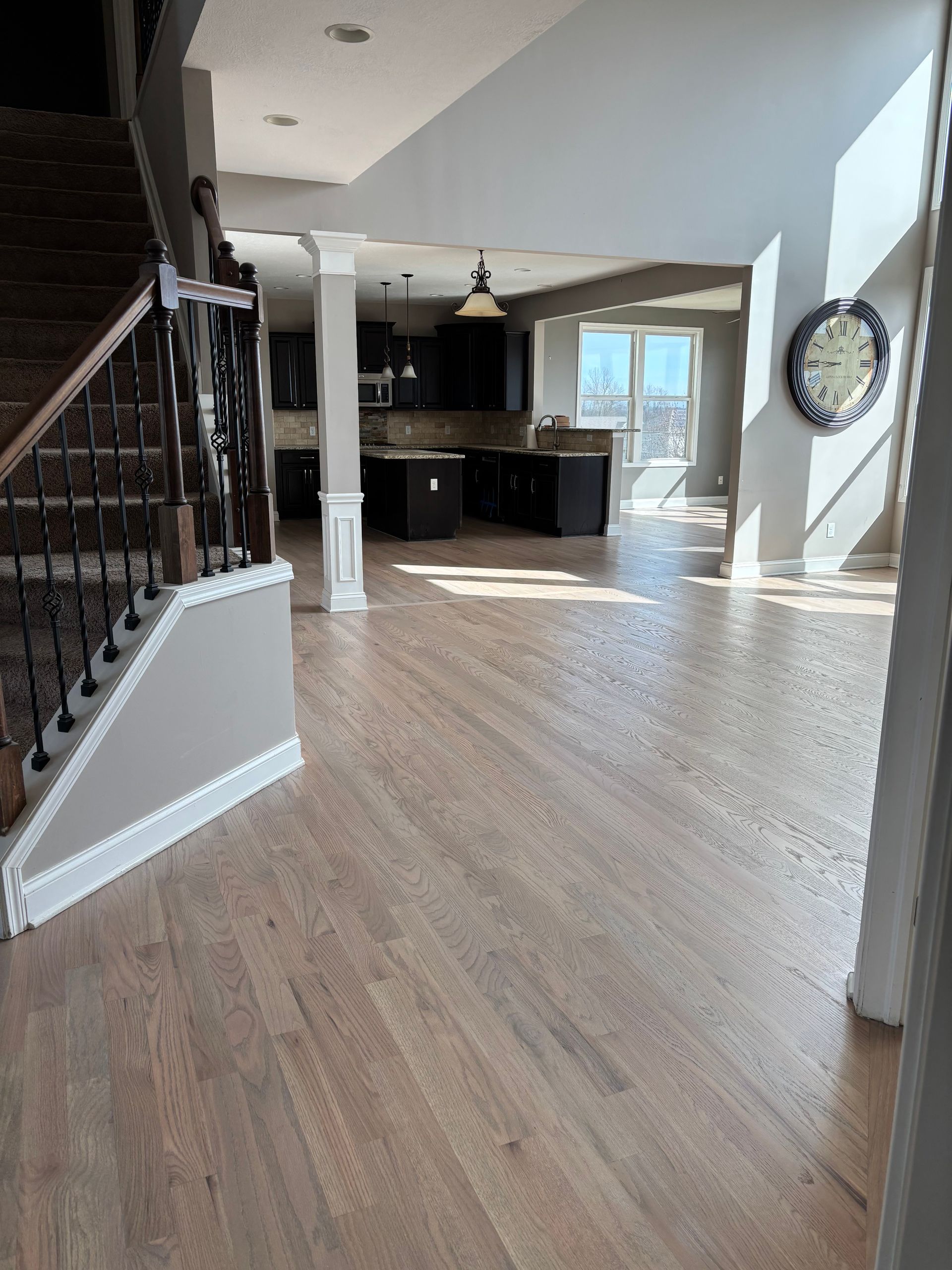 Bright open living room with hardwood floors, staircase, and dark kitchen cabinets by a sunlit doorway