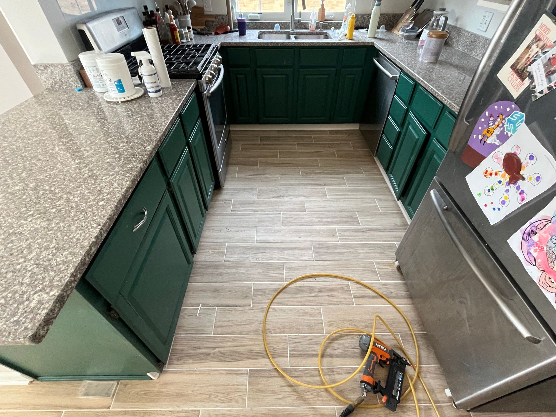 Kitchen with green cabinets, gray tile floor, and a power tool on the floor by the refrigerator.