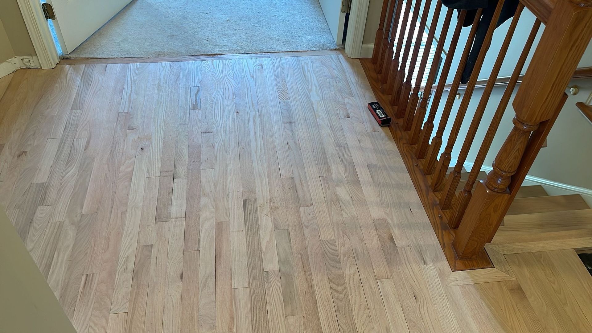 Light wood flooring in a hallway next to a staircase with wooden handrails.