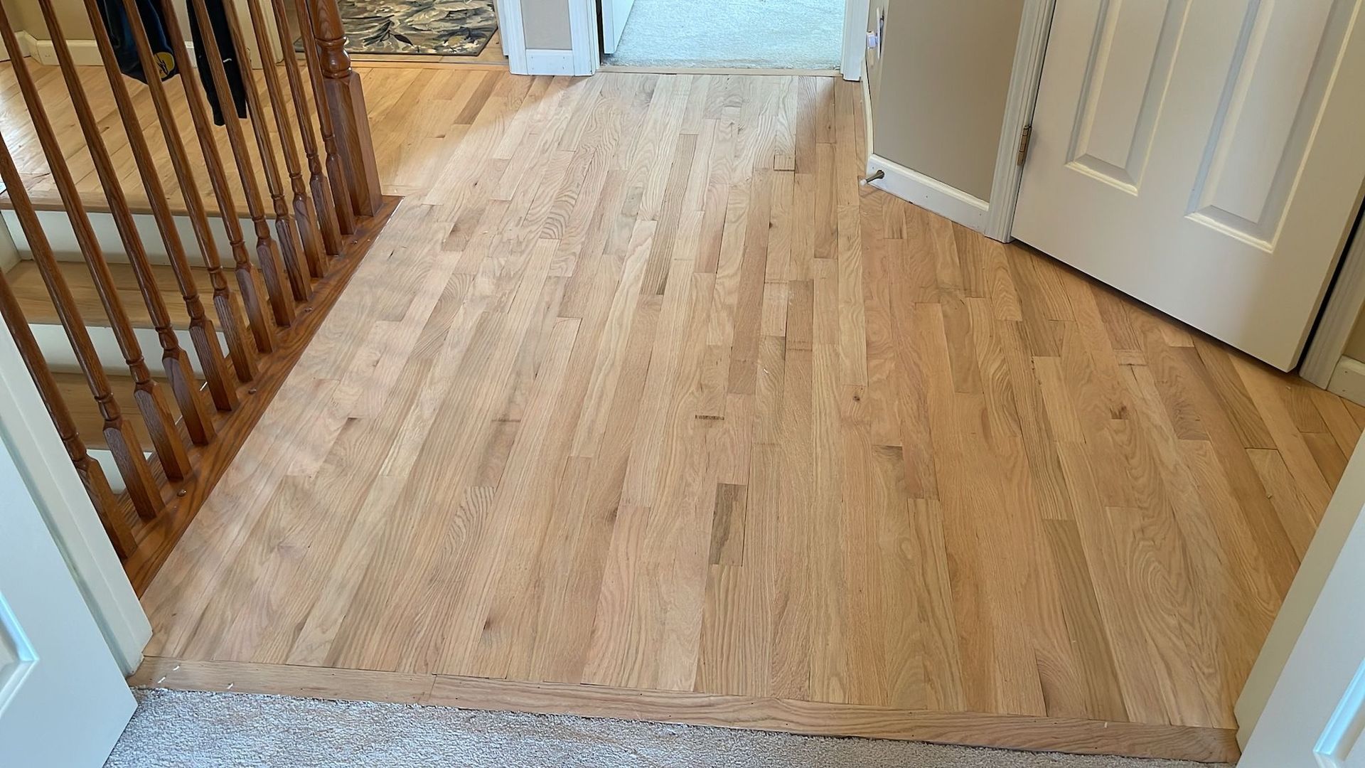 Hardwood flooring transition to carpeted hallway. Door on the right, wood banister on the left.