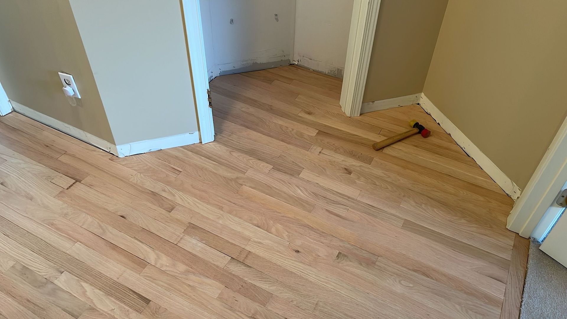 Hardwood floors being installed near closet. Hammer lies on the floor. Beige walls and white trim.