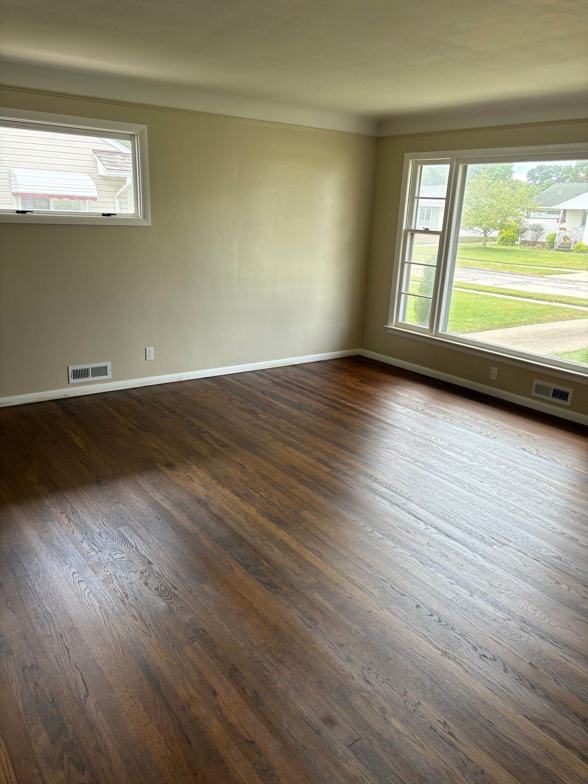 Empty living room with dark wood floors and light walls. Two windows provide natural light.