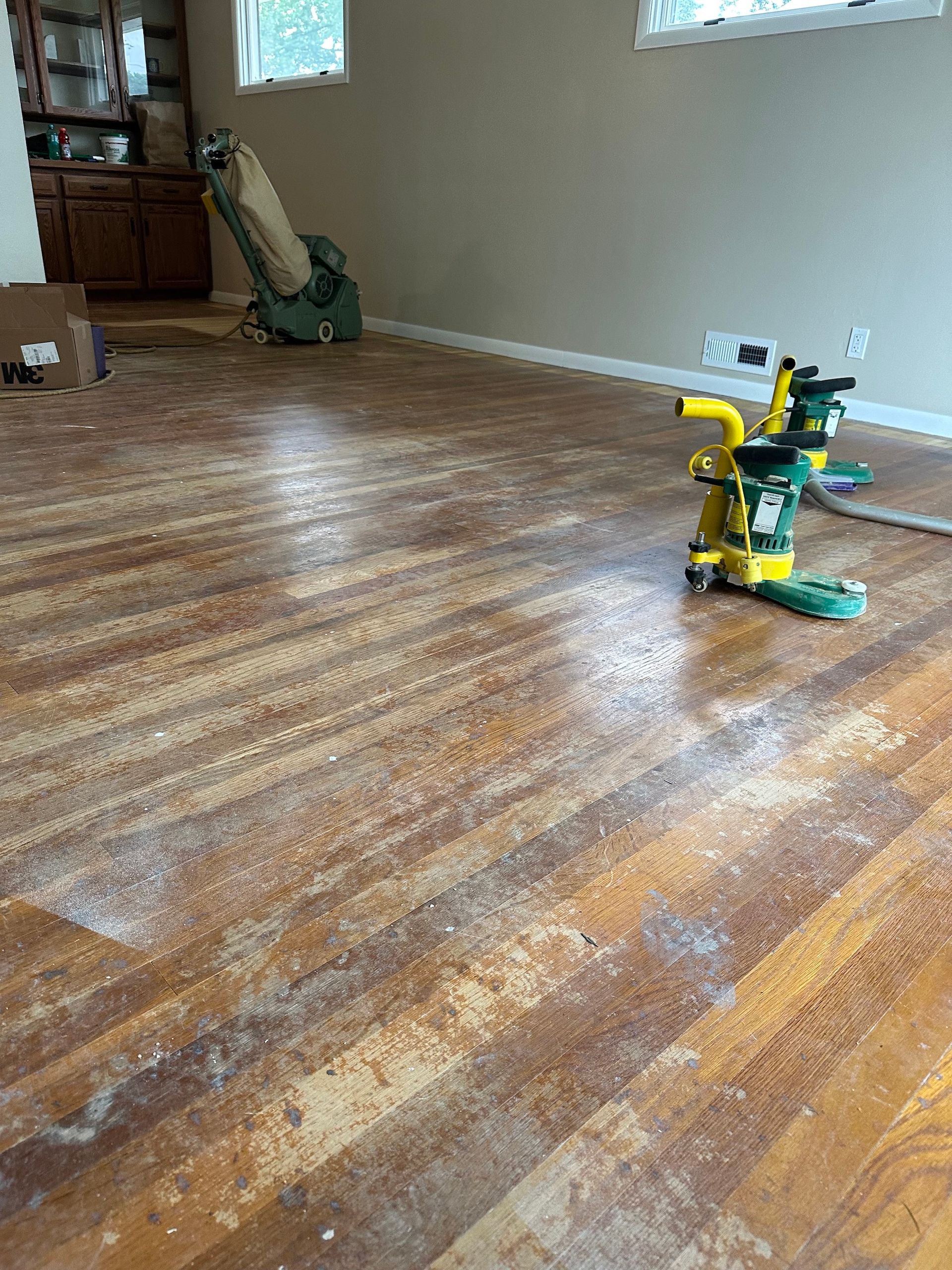 Wooden floor being sanded, with sanding machines visible. Dust and debris cover the floor.