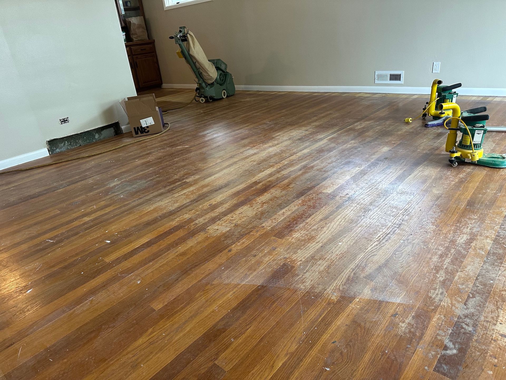 Wooden floor being sanded; two yellow sanders and dust present in a room.