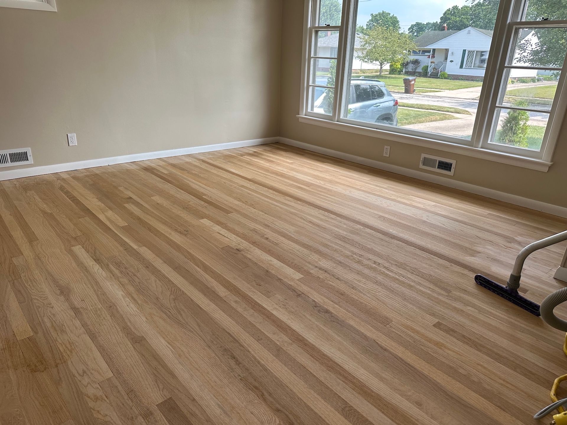 Newly refinished hardwood floor in a room with windows, beige walls, and white trim.