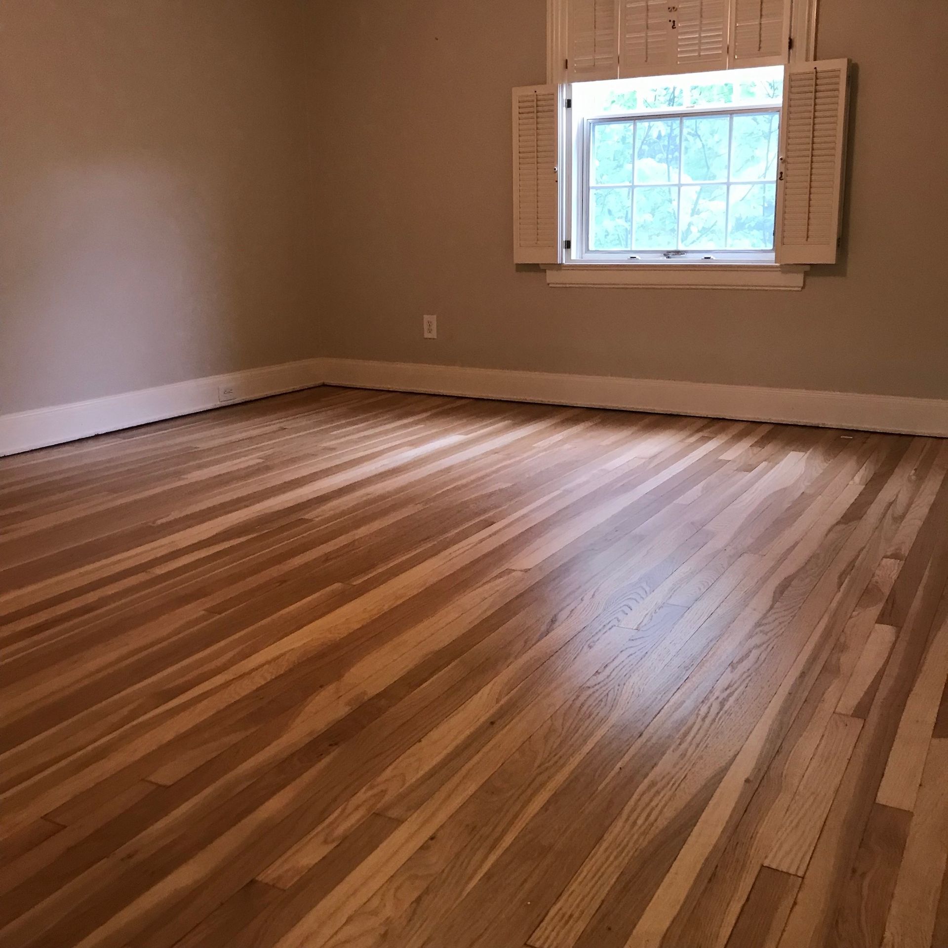 Empty room with wood floors and a window with shutters. Beige walls and natural light.