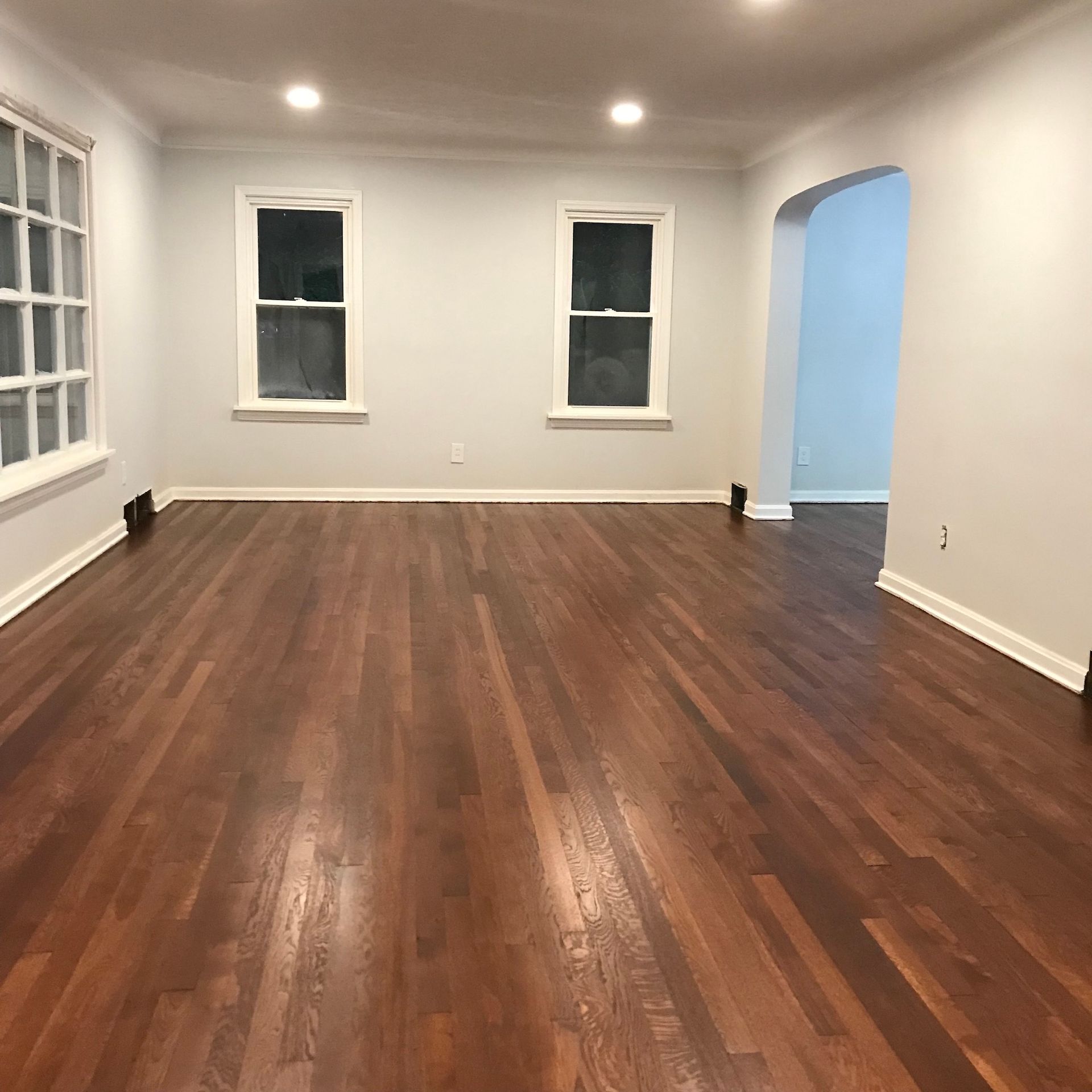 Empty room with hardwood floor, white walls, windows, and an arched doorway.