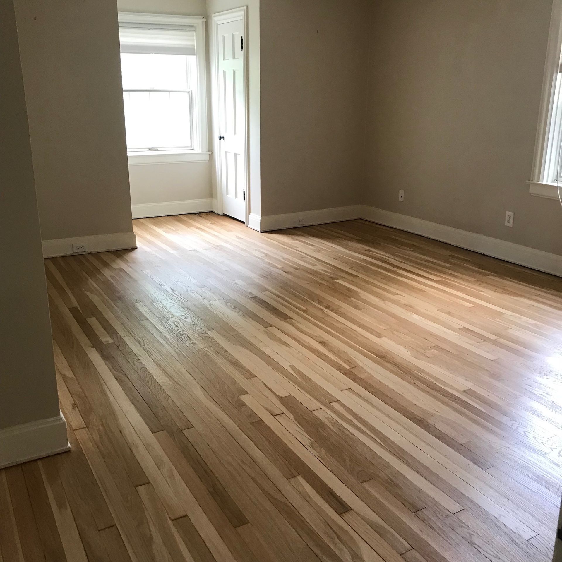Empty room with newly refinished hardwood floors, windows, and beige walls.