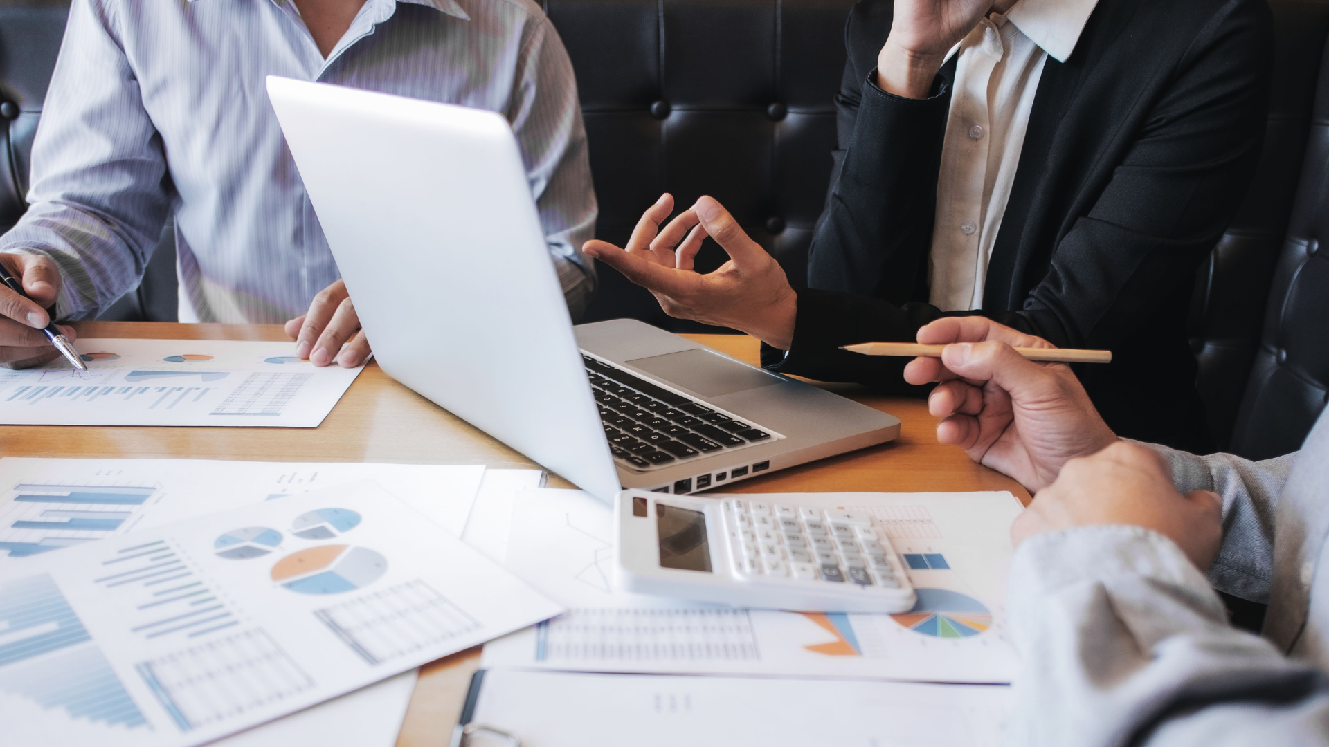 People in business attire are at a table with a laptop, charts, and a calculator, discussing data.