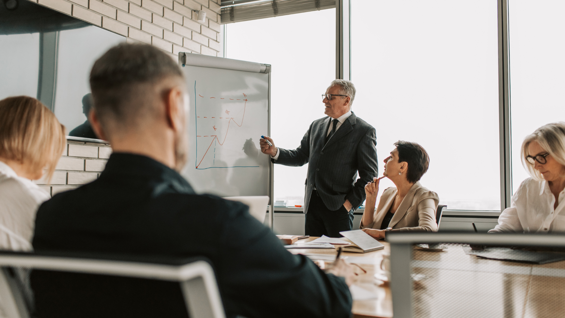 Business presentation: man points at whiteboard with graph, facing seated colleagues in a conference room.