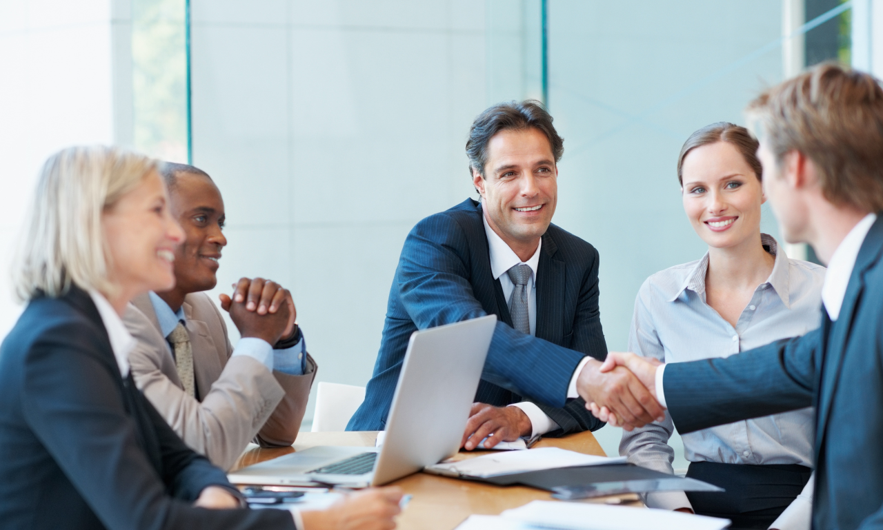 A group of business people are sitting around a table shaking hands.